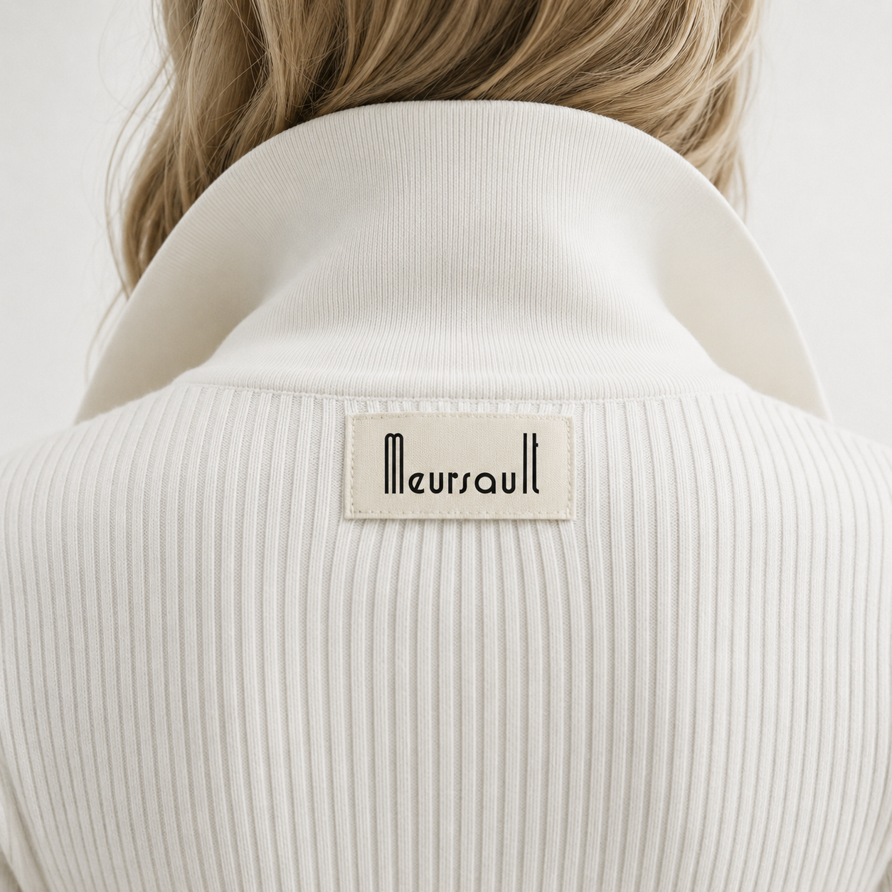 Back view of a person wearing a white ribbed turtleneck sweater with a label that says "Meursault."