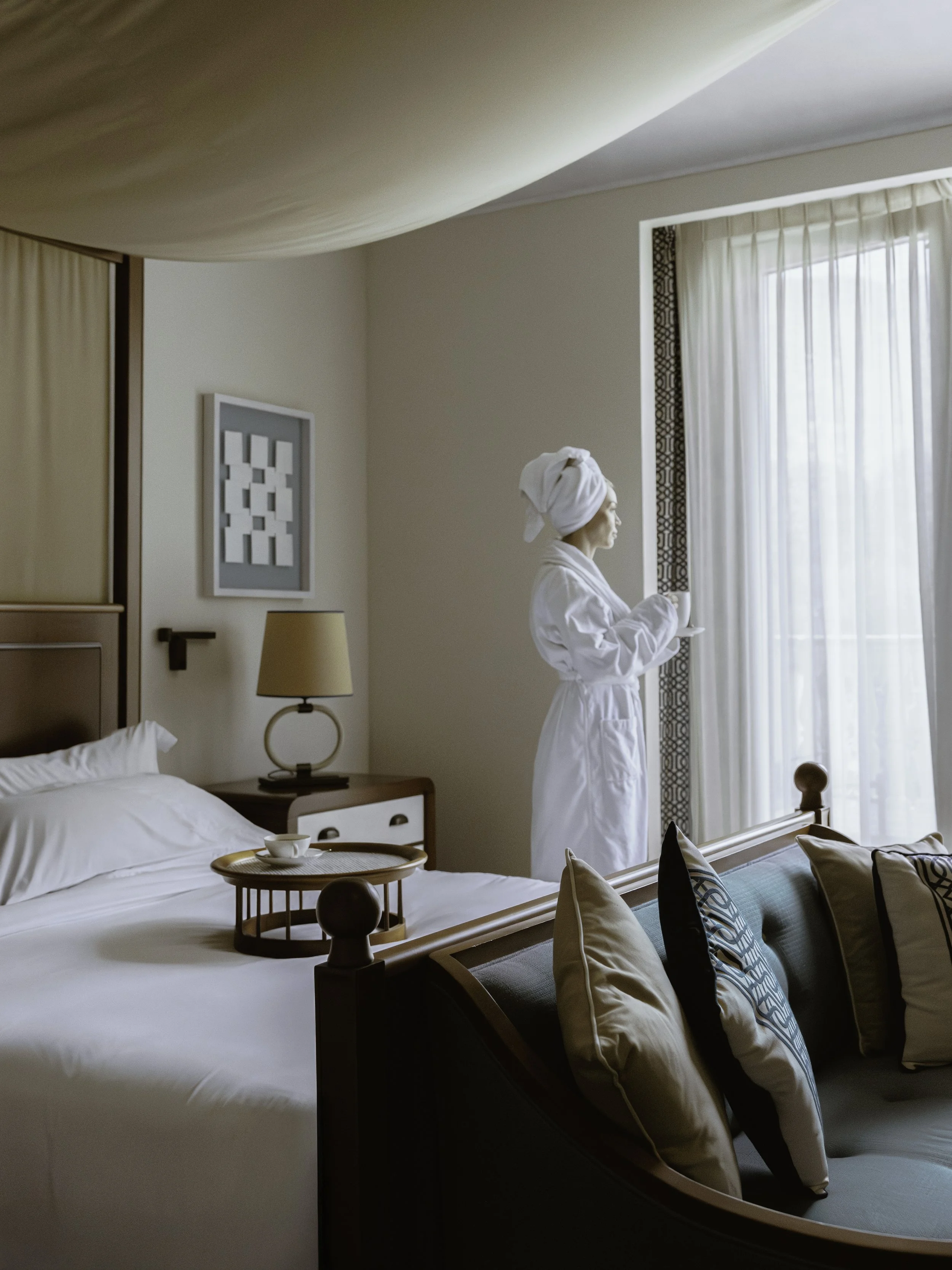 A woman wearing a white robe and towel wrapped around her head stands in a hotel room, looking out the window. The room has a bed with white bedding, a wooden bedside table with a tray holding a teacup, beige walls, a framed geometric art piece, and 