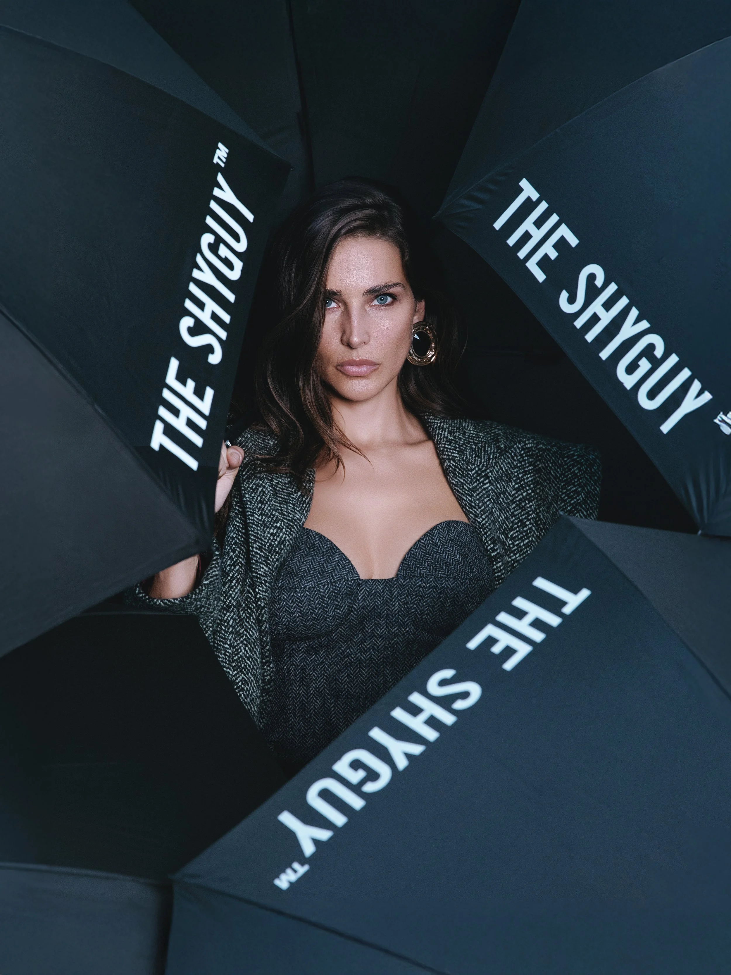 A woman with dark hair and blue eyes holding black umbrellas with white text that reads 'THE SHYGUY' and 'SHYGUY TM'.