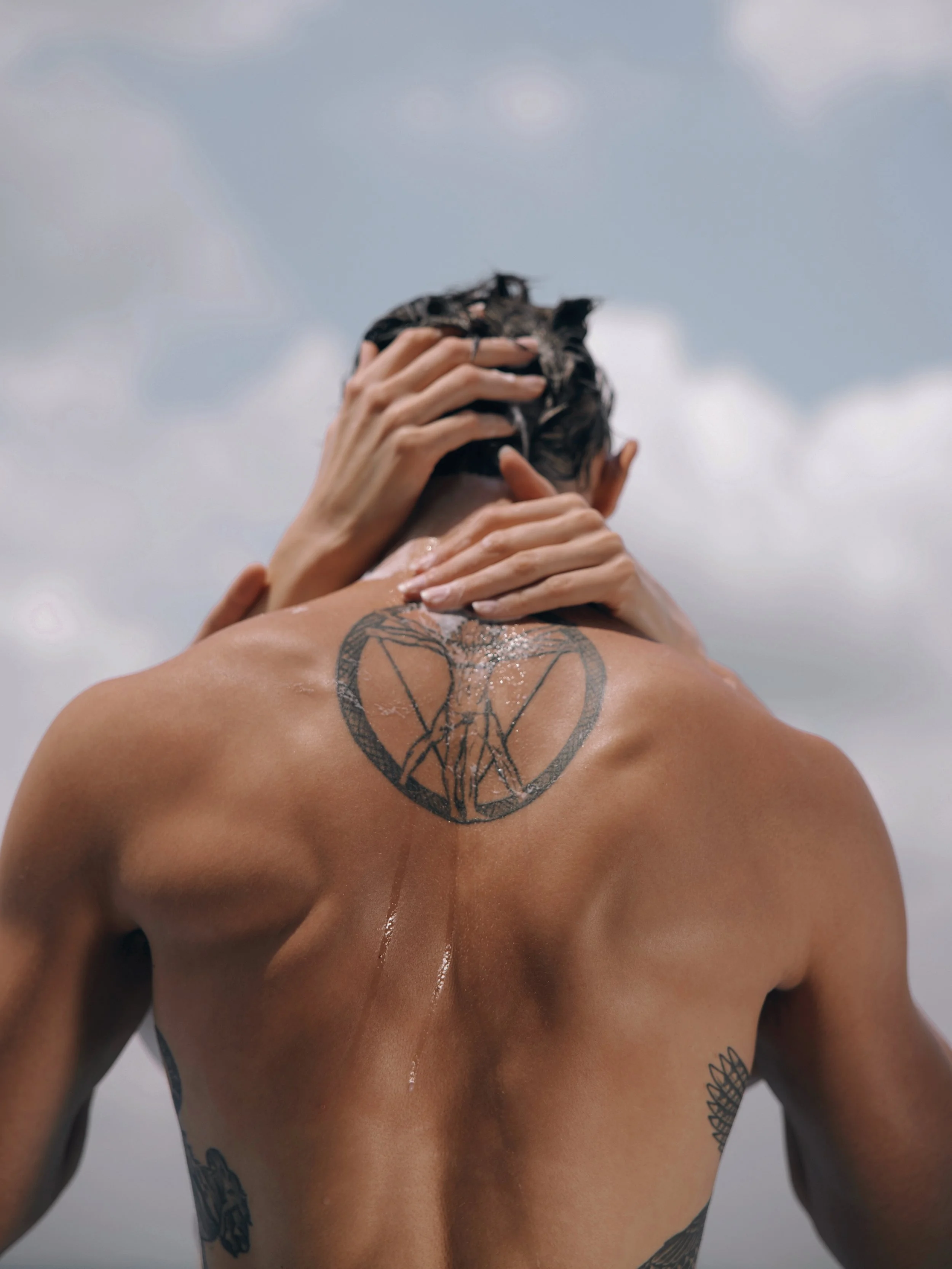 A person with wet hair and tattoos on their back, holding their head with both hands against a cloudy sky.