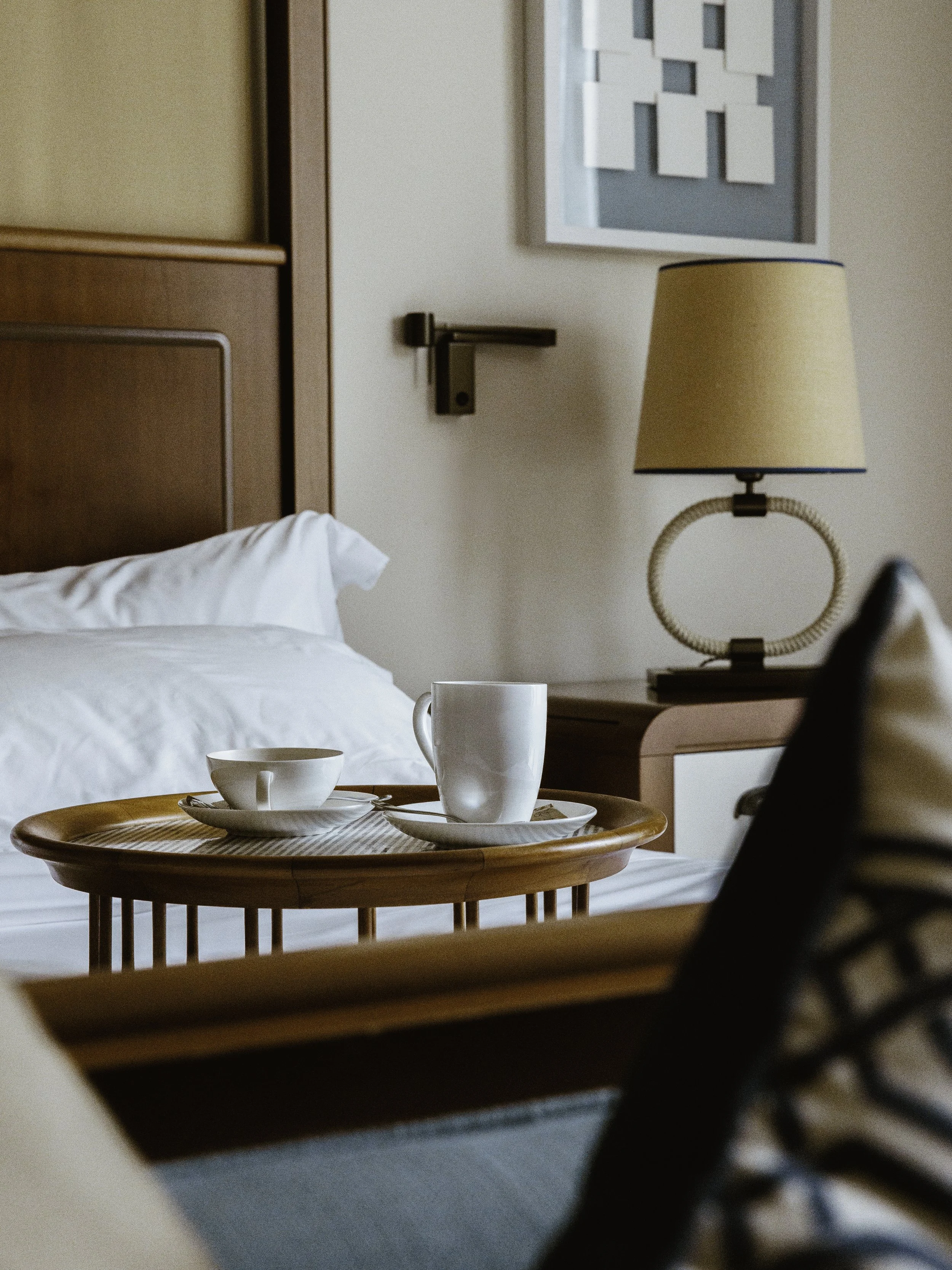 A hotel room with a bed, nightstand, lamp, and tea cups on a wooden tray, with wall art and a door handle visible in the background.
