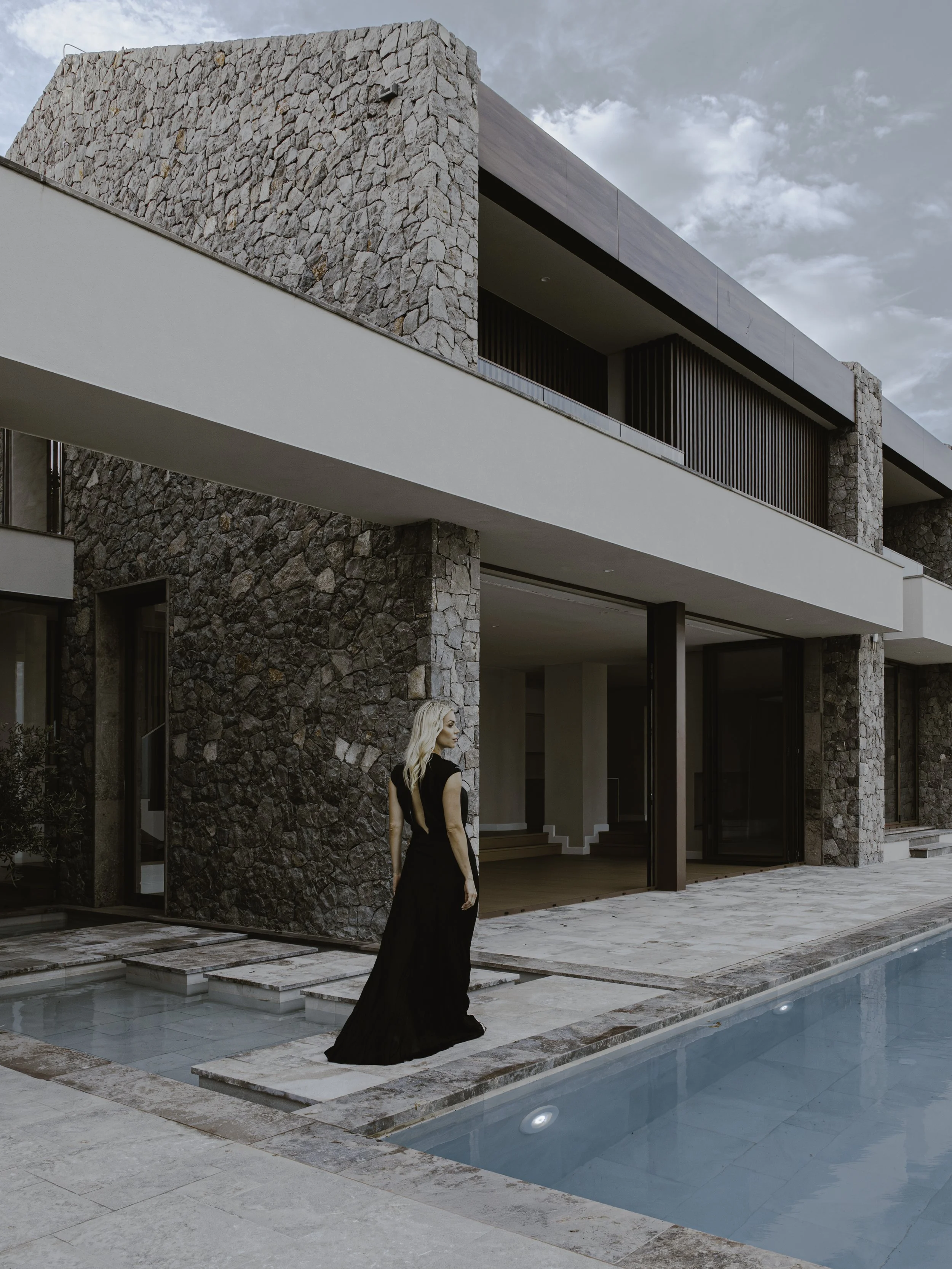 A woman in a long black gown walking near a modern house with stone walls and a swimming pool.