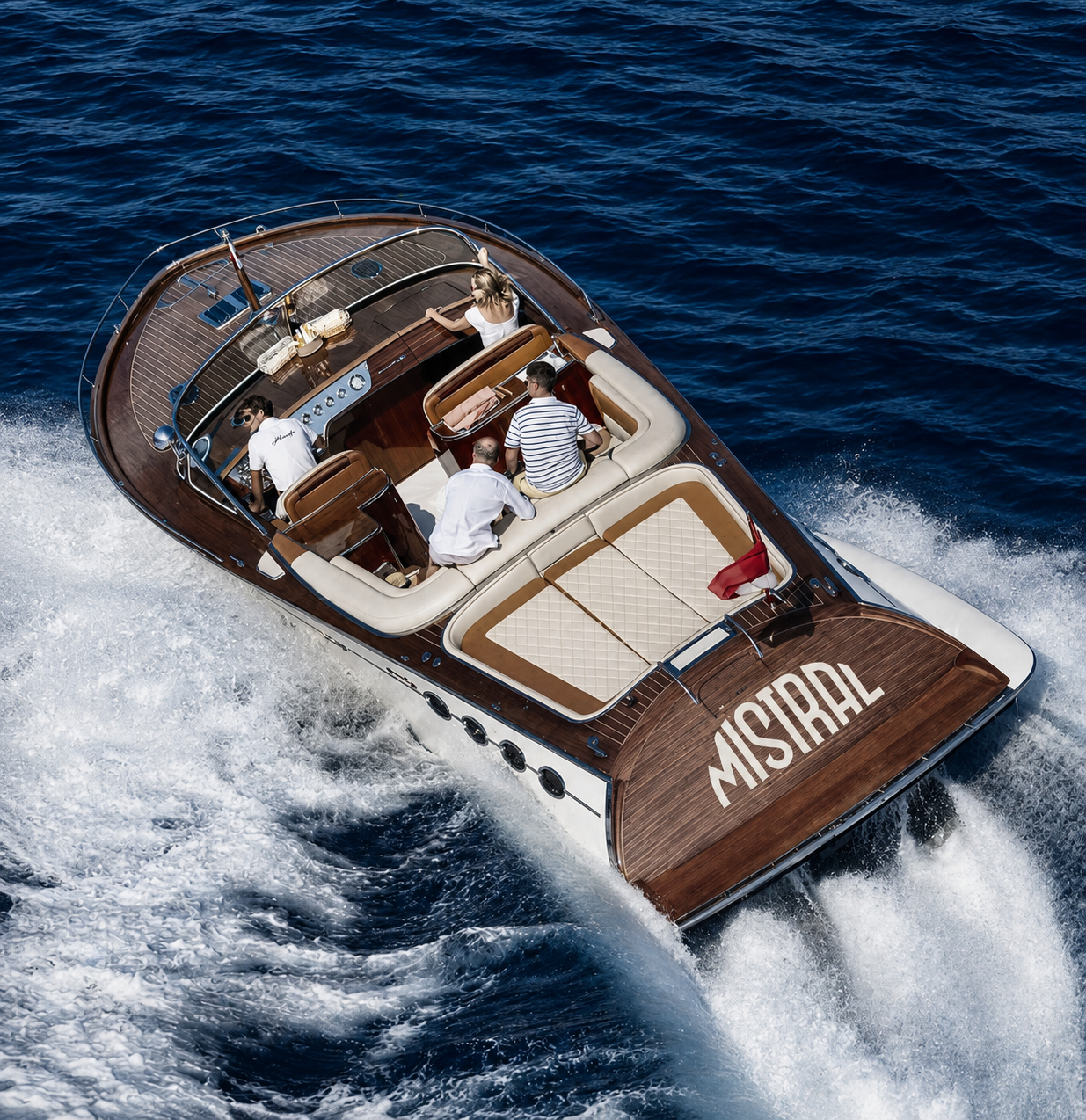 A luxury motorboat with four people on board cruising on the ocean. The boat has natural wood and white seating, and the name 'MISIRAL' on the stern. The water is deep blue and choppy, with white foamy waves.