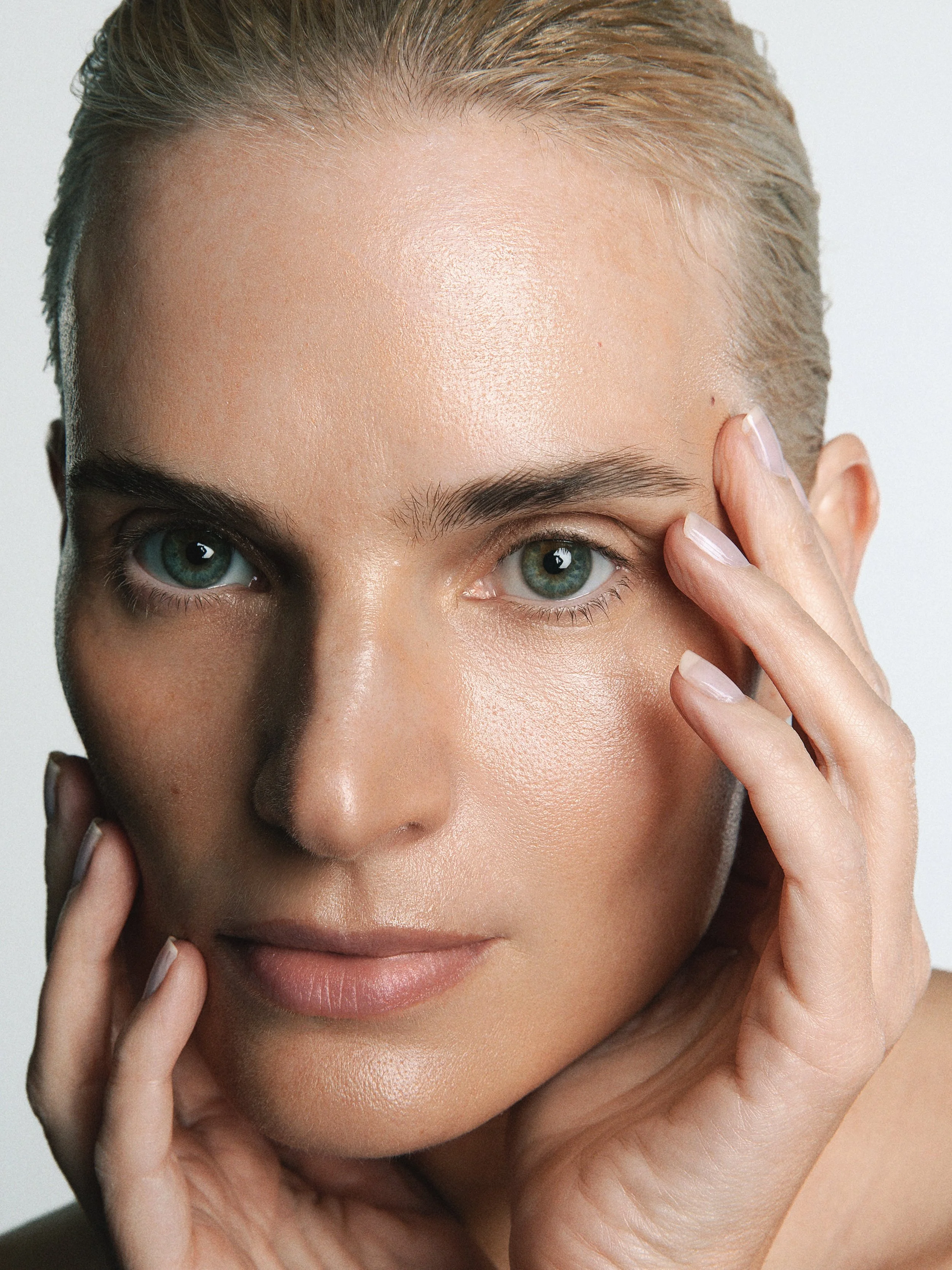 Close-up of a woman's face with light makeup, touching her forehead and cheek gently with her fingers, against a plain background.