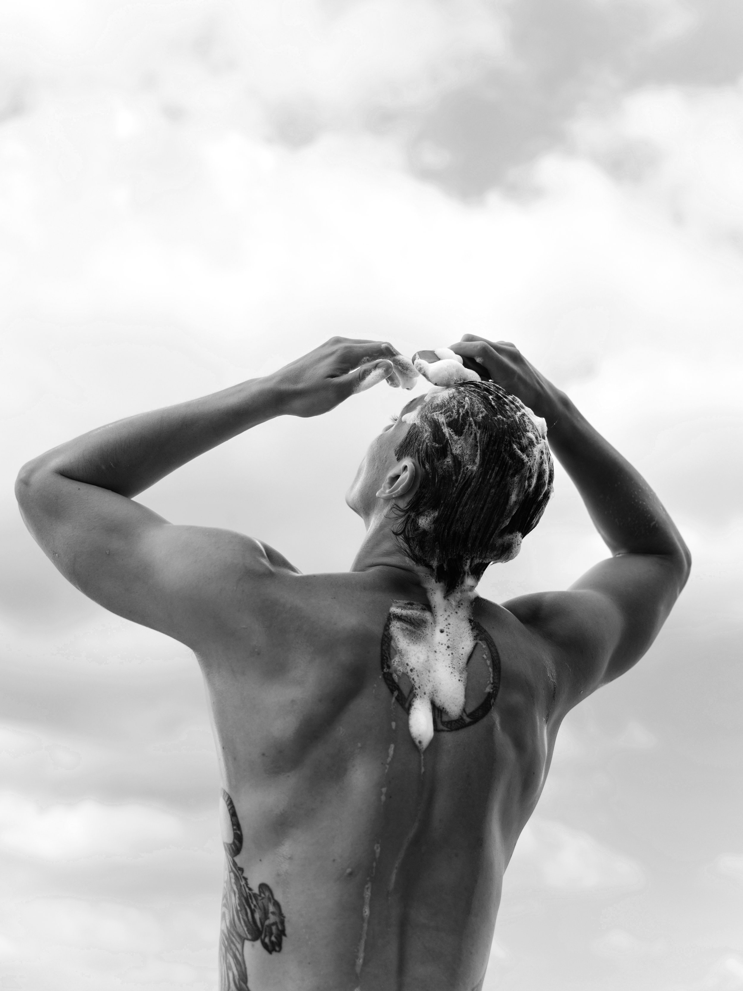 A person washing their hair with soap and water in an outdoor setting, with clouds in the sky in the background.