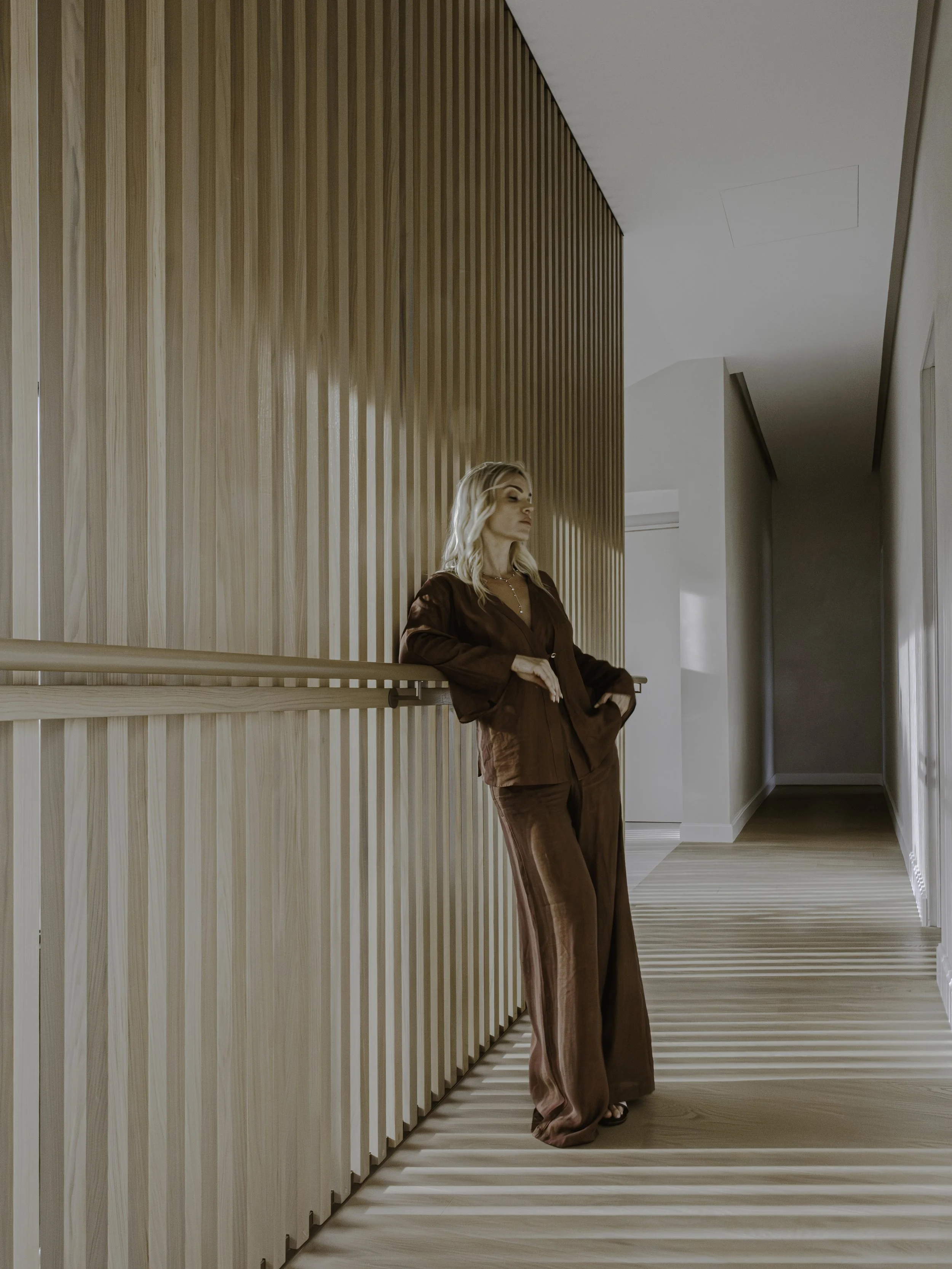 A woman with blonde hair leaning against a wooden-paneled wall in a modern interior space, wearing a brown satin suit with wide-leg pants and a matching top, with her eyes closed and hands resting on her hips.