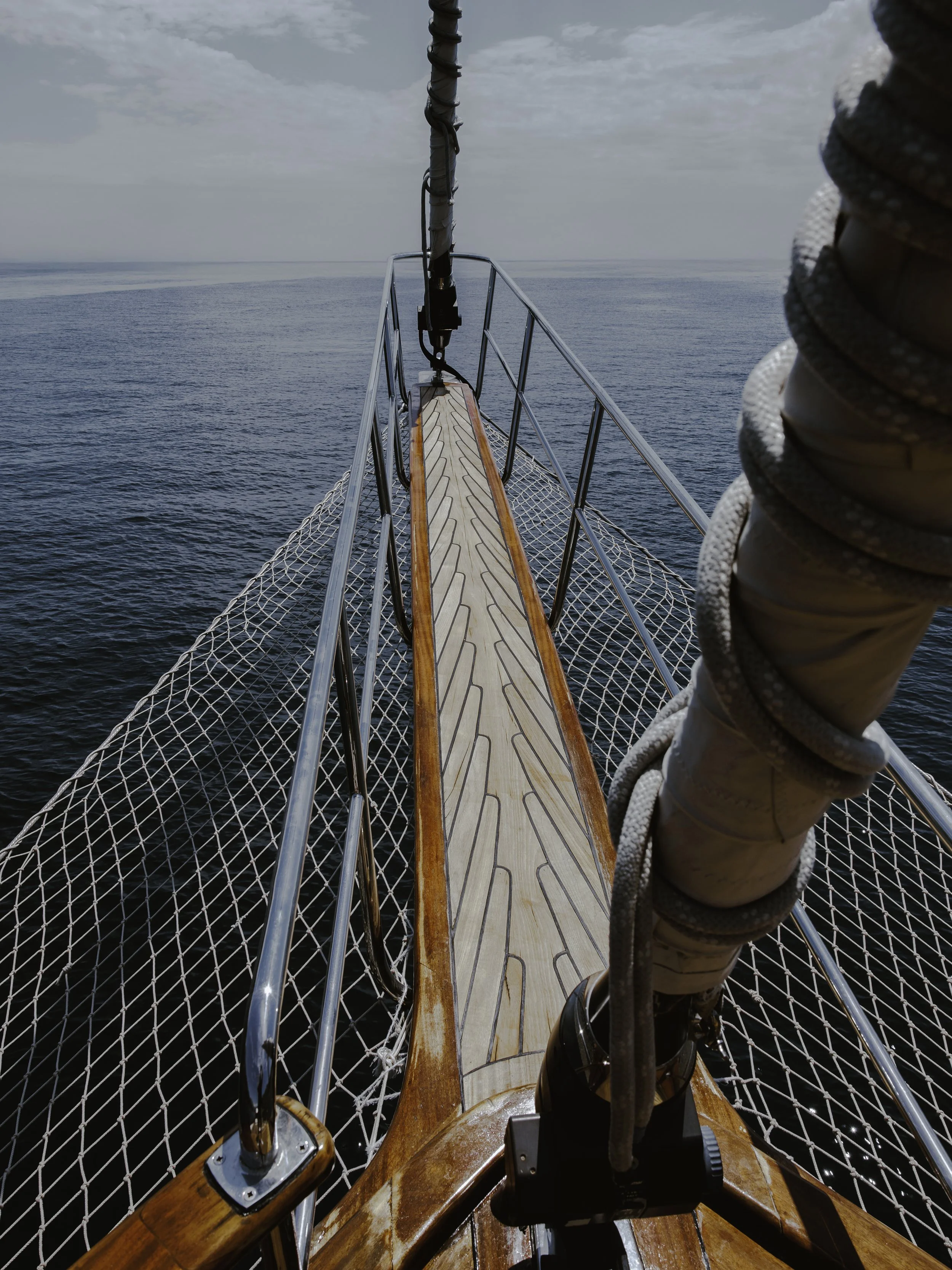 View from the bow of a boat with a wooden walkway, metal railings, and a rope bundle on the right side, overlooking the open ocean under cloudy sky.