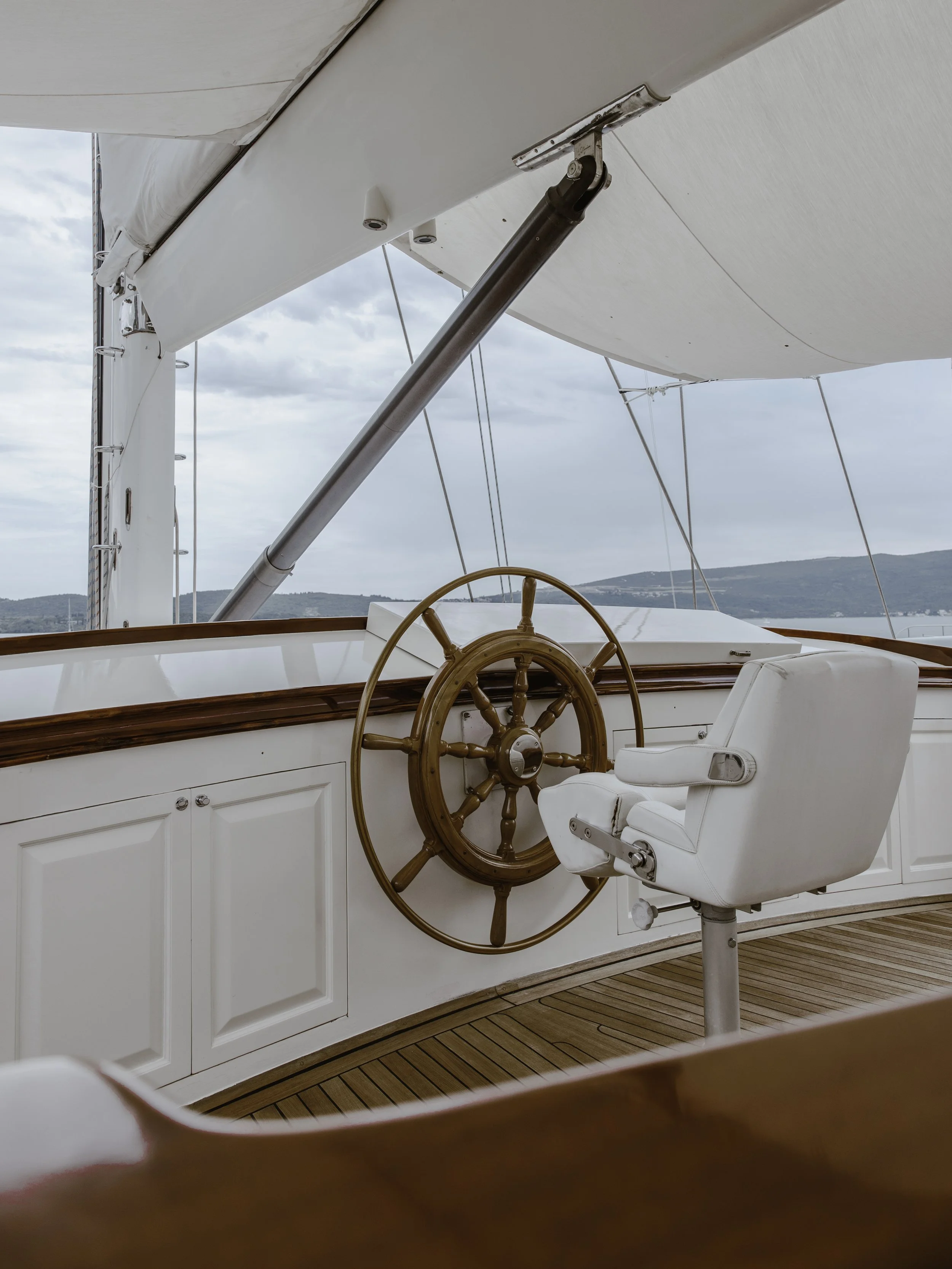 View of a boat's helm with a wooden steering wheel and white captain's chair, under a large white canopy, with water and hills in the background.
