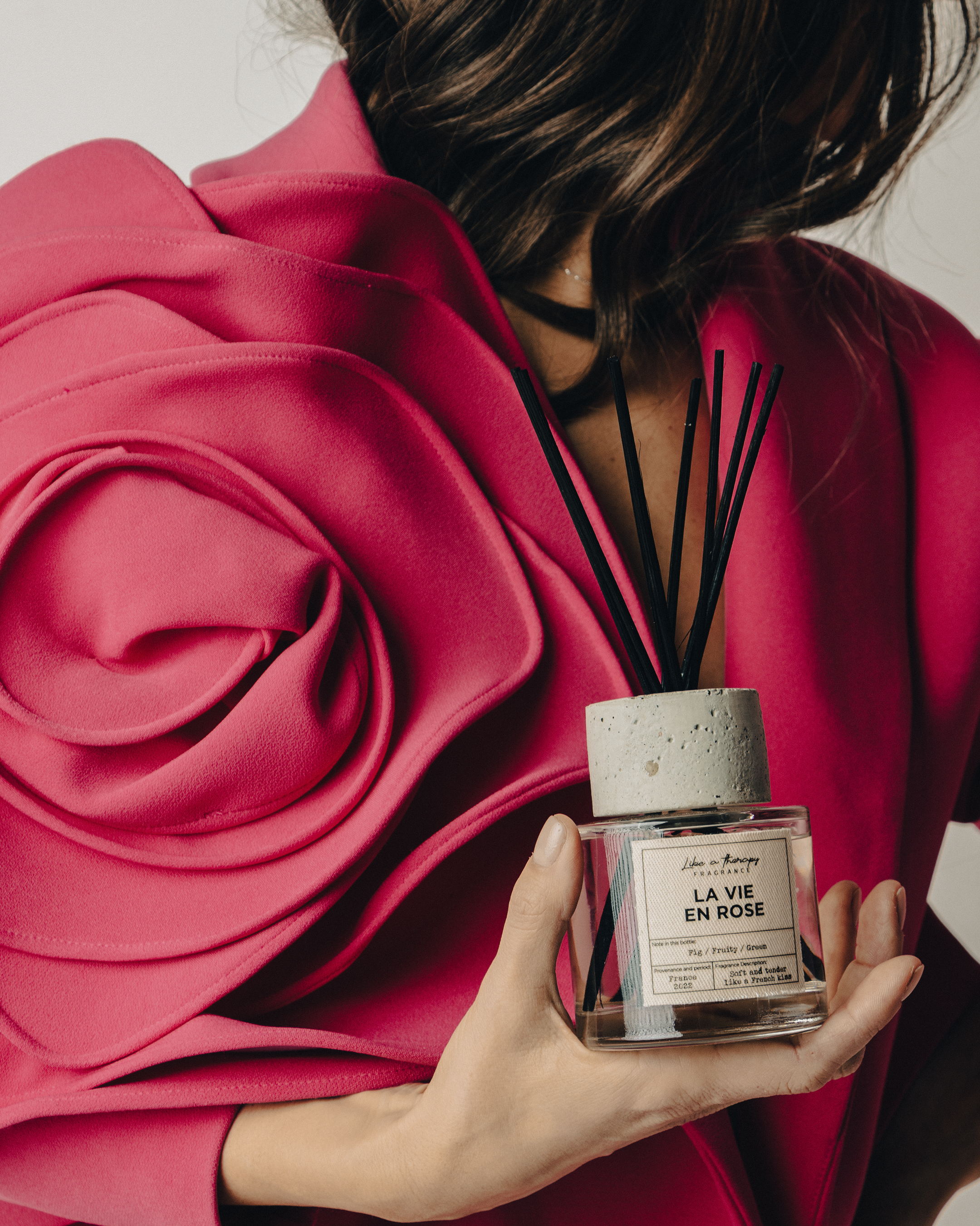 Person wearing a pink blazer holding a small glass fragrance bottle with black reed sticks inside. The label on the bottle reads "LA VIE EN ROSE" and includes additional description. The person’s face is not visible in the image.