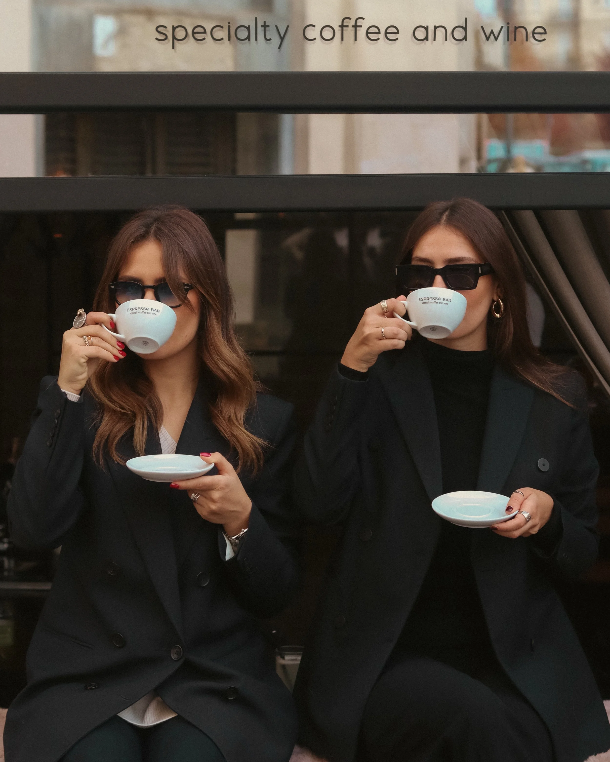 Two women wearing black blazers and sunglasses are drinking coffee from white cups with the label "Espresso Bar" and a saucer, sitting side by side in front of a black window. Their faces are partially obscured by the cups.