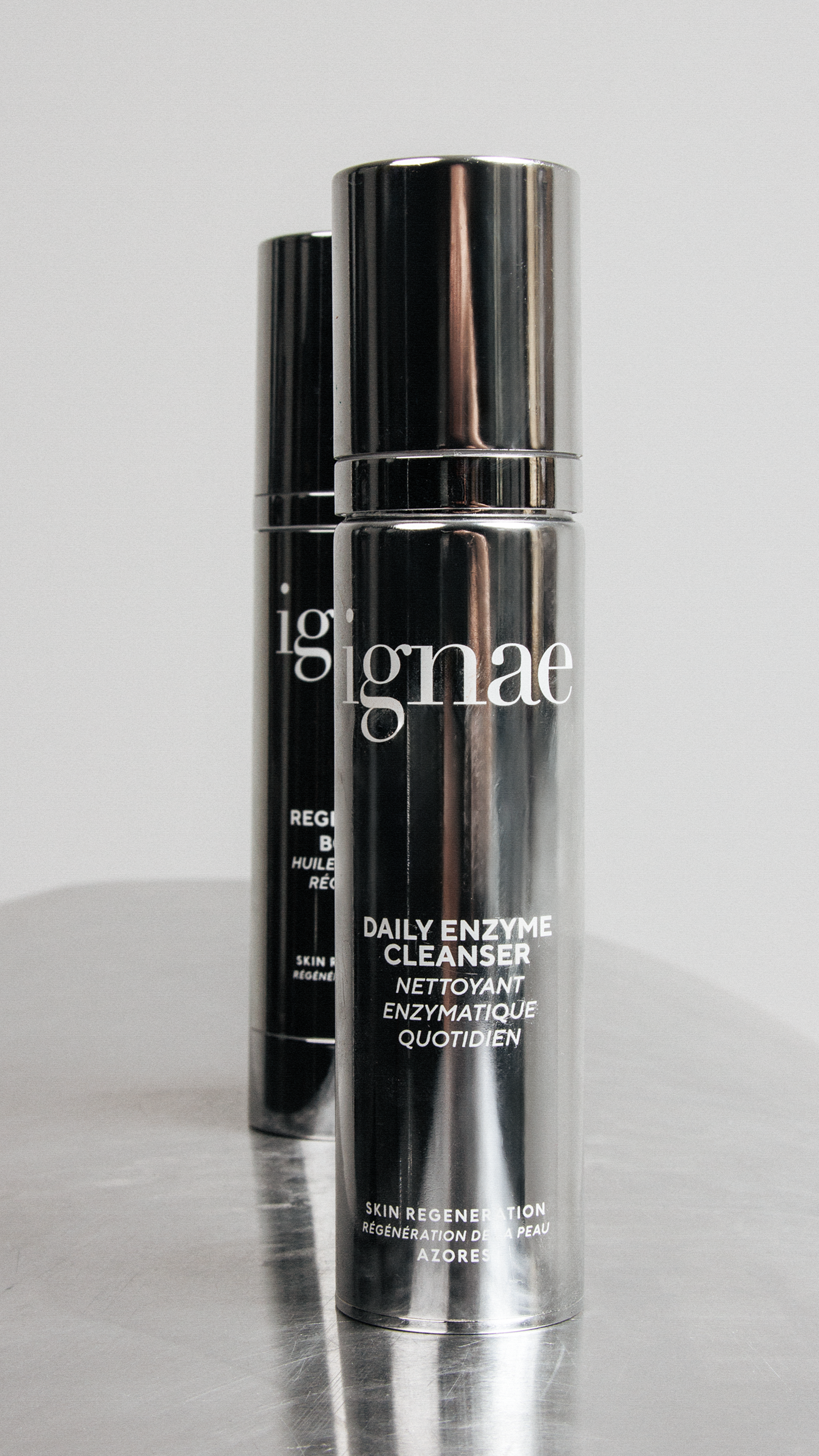 Two sleek, black cylindrical bottles of skincare product, labeled 'Daily Enzyme Cleanser,' with silver caps, standing on a reflective surface.