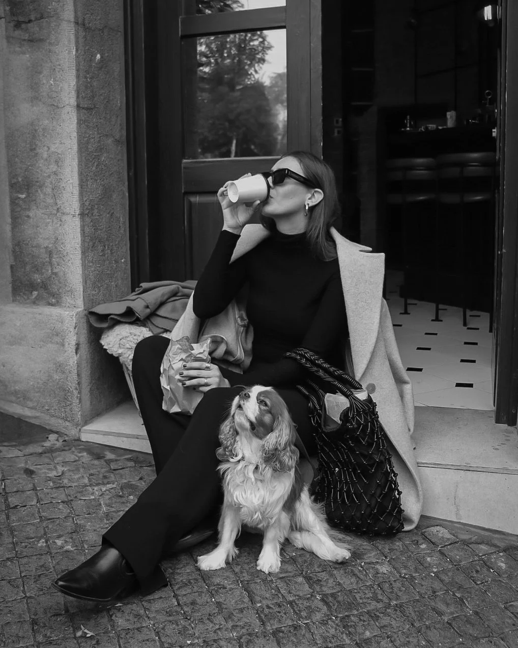 A woman with shoulder-length hair, wearing sunglasses, a black turtleneck, and black pants, is sitting outside a cafe or restaurant, drinking from a paper cup. She has a coat draped over her shoulders and is sitting on a small stool with a dog sittin