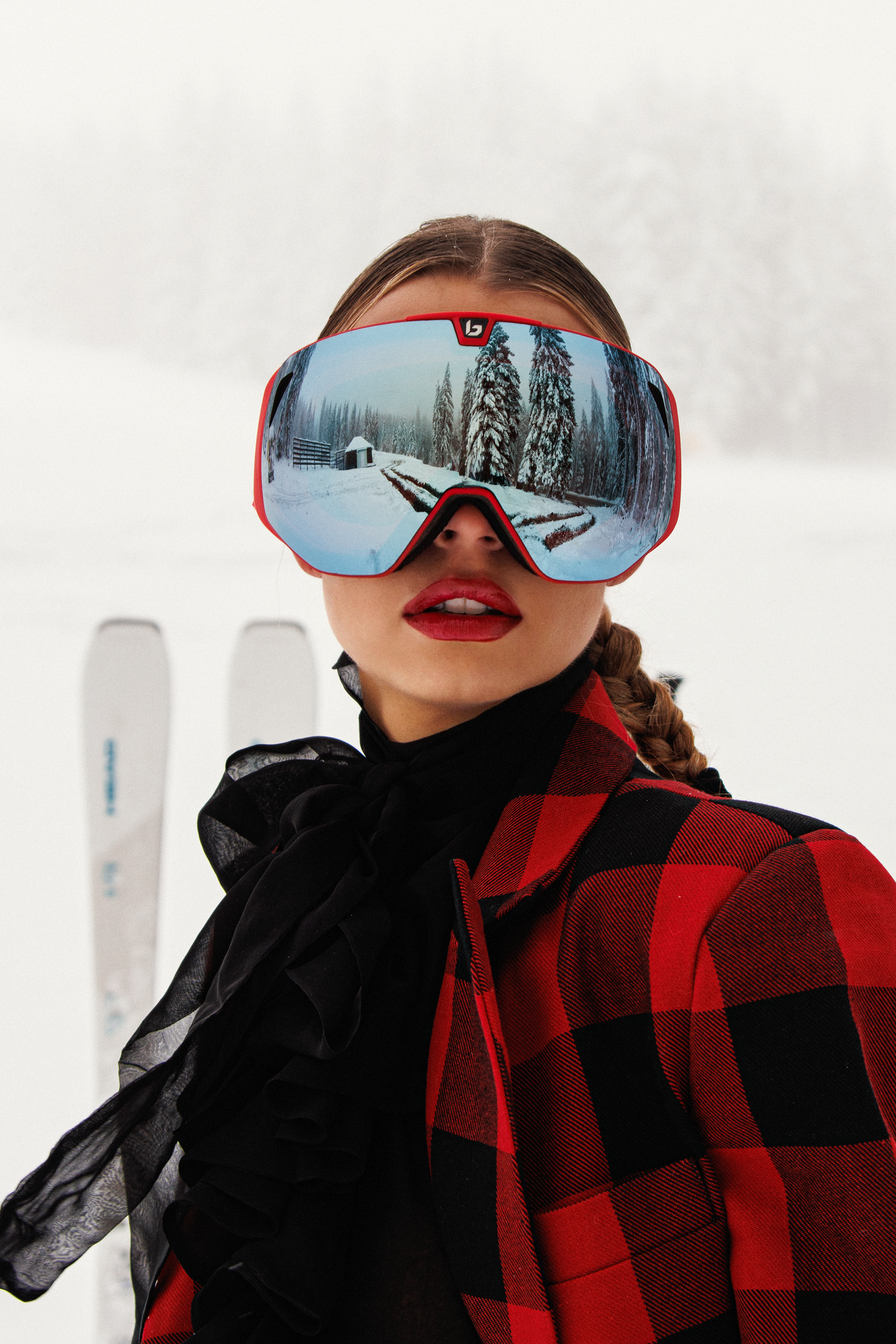 A woman wearing large ski goggles reflecting snowy trees and a cabin, dressed in a red and black plaid jacket and black gloves, with skiing equipment in the background.