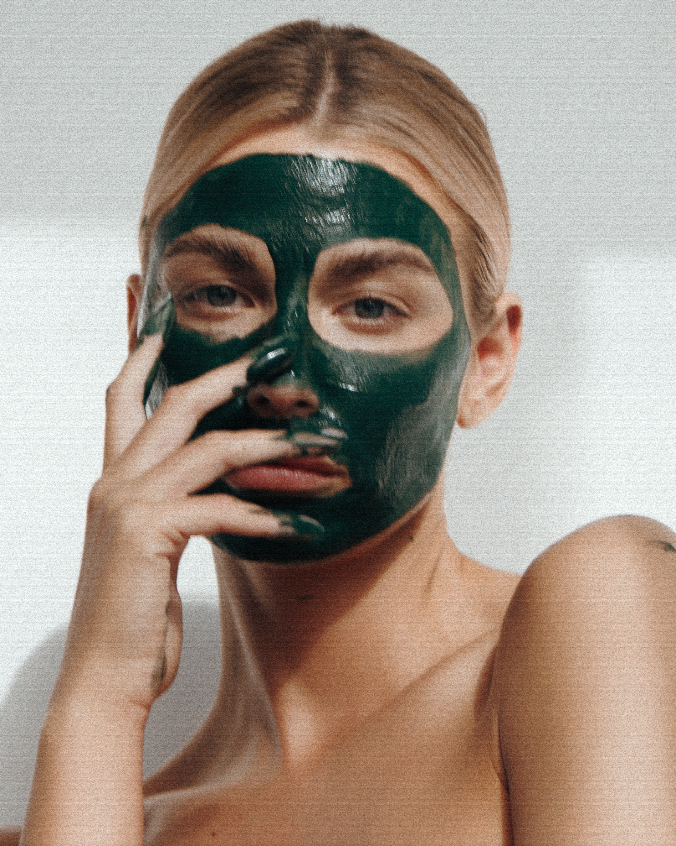 A person with blonde hair and light skin is holding a green face mask close to their face, partially covering it. The person appears to be topless and is standing against a plain, light-colored background.