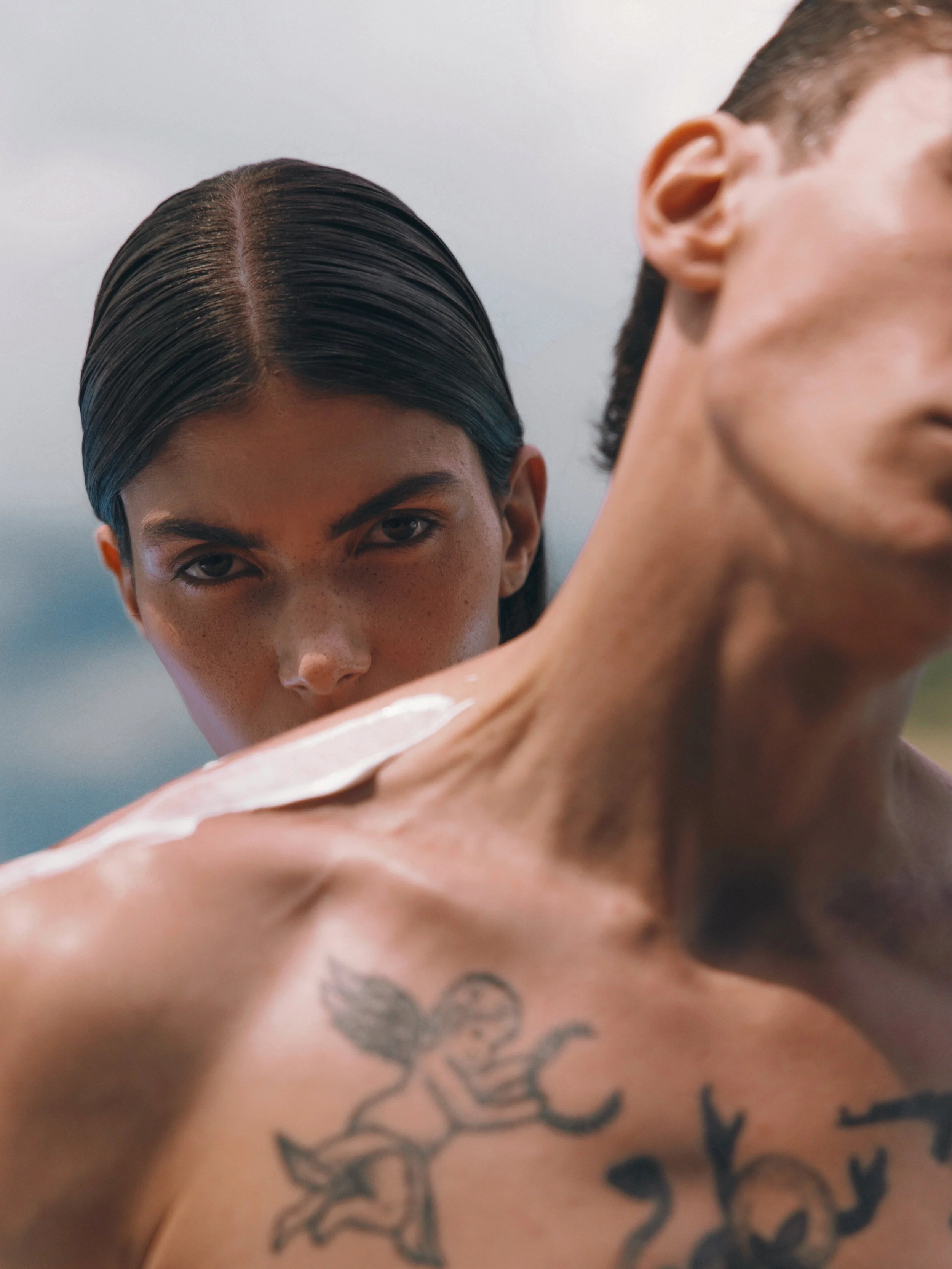 Close-up of two people, one with dark, slicked-back hair and the other with short hair and a visible tattoo on their shoulder, outdoors with a cloudy sky in the background.