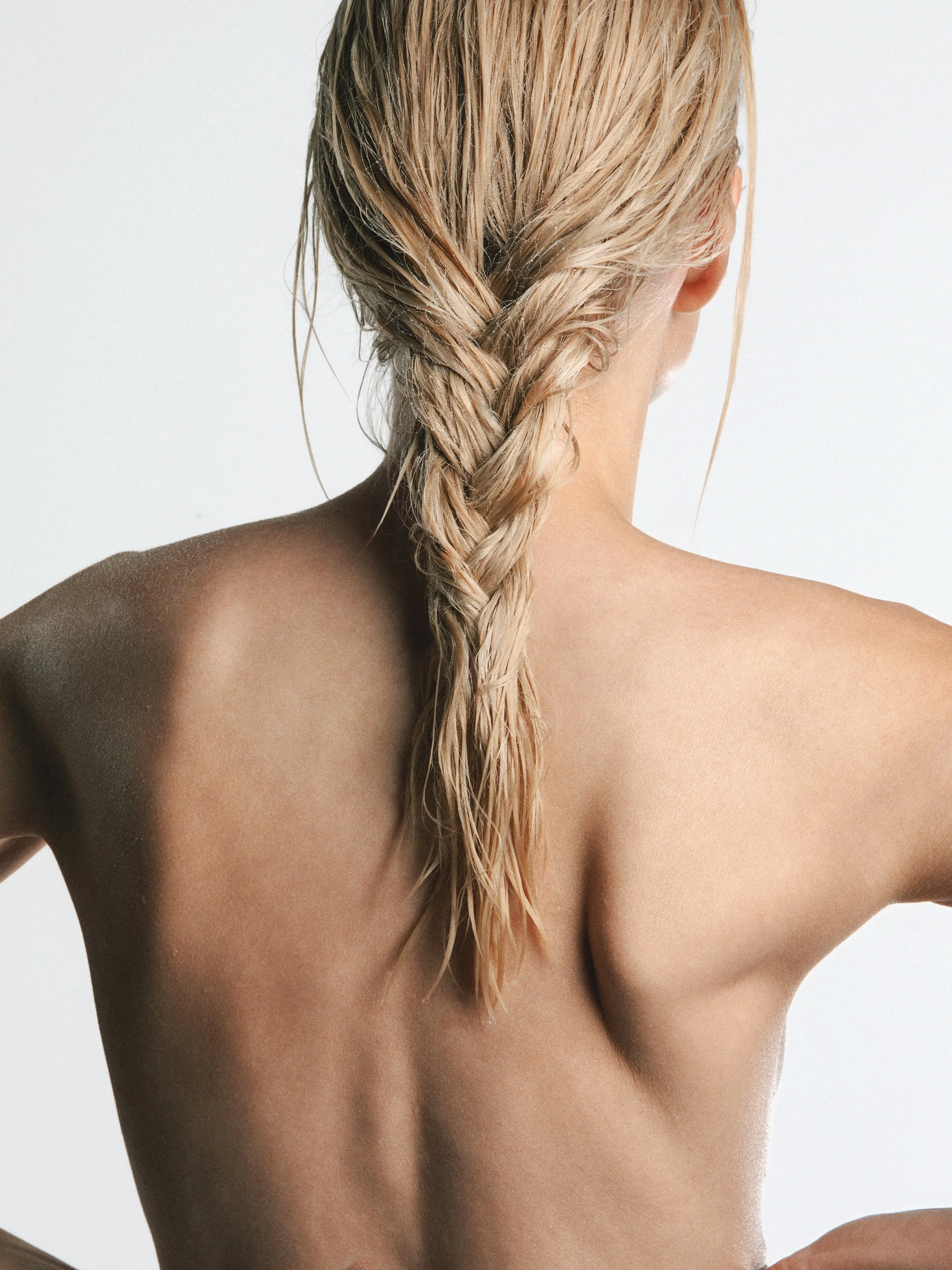 Back view of a woman with blonde hair in a braid, with bare shoulders and partially visible arm against a plain background.