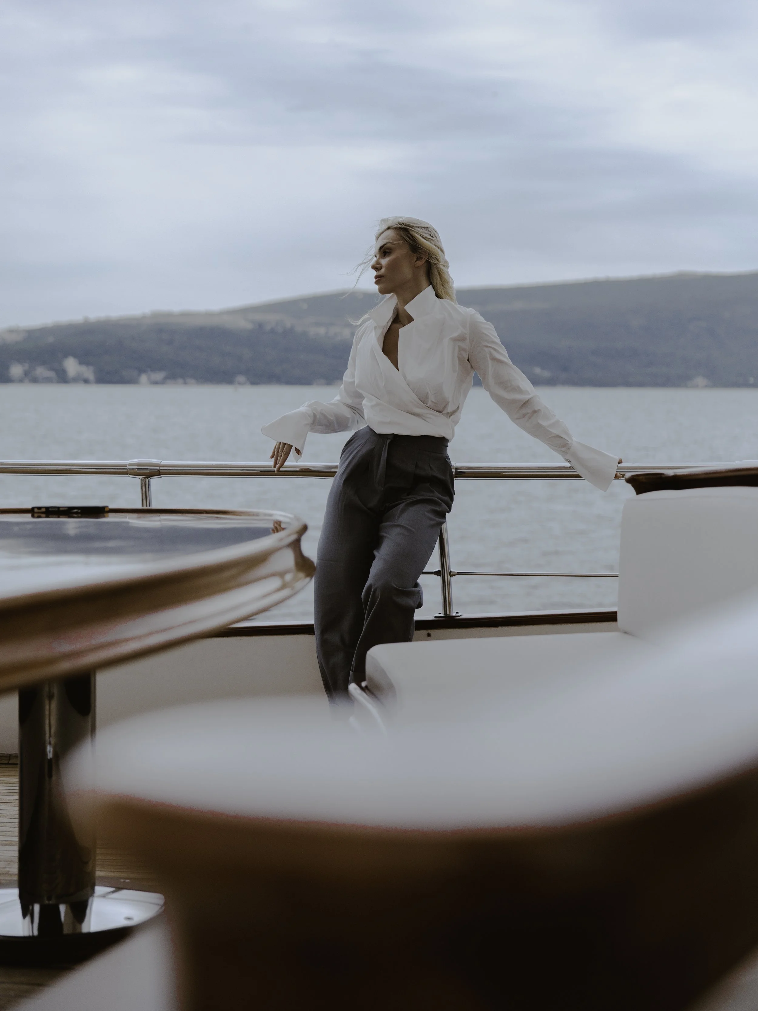 A woman with blonde hair in a white blouse and dark pants stands on a boat deck, holding onto a railing, with a body of water and a hilly landscape in the background. The scene is under an overcast sky.