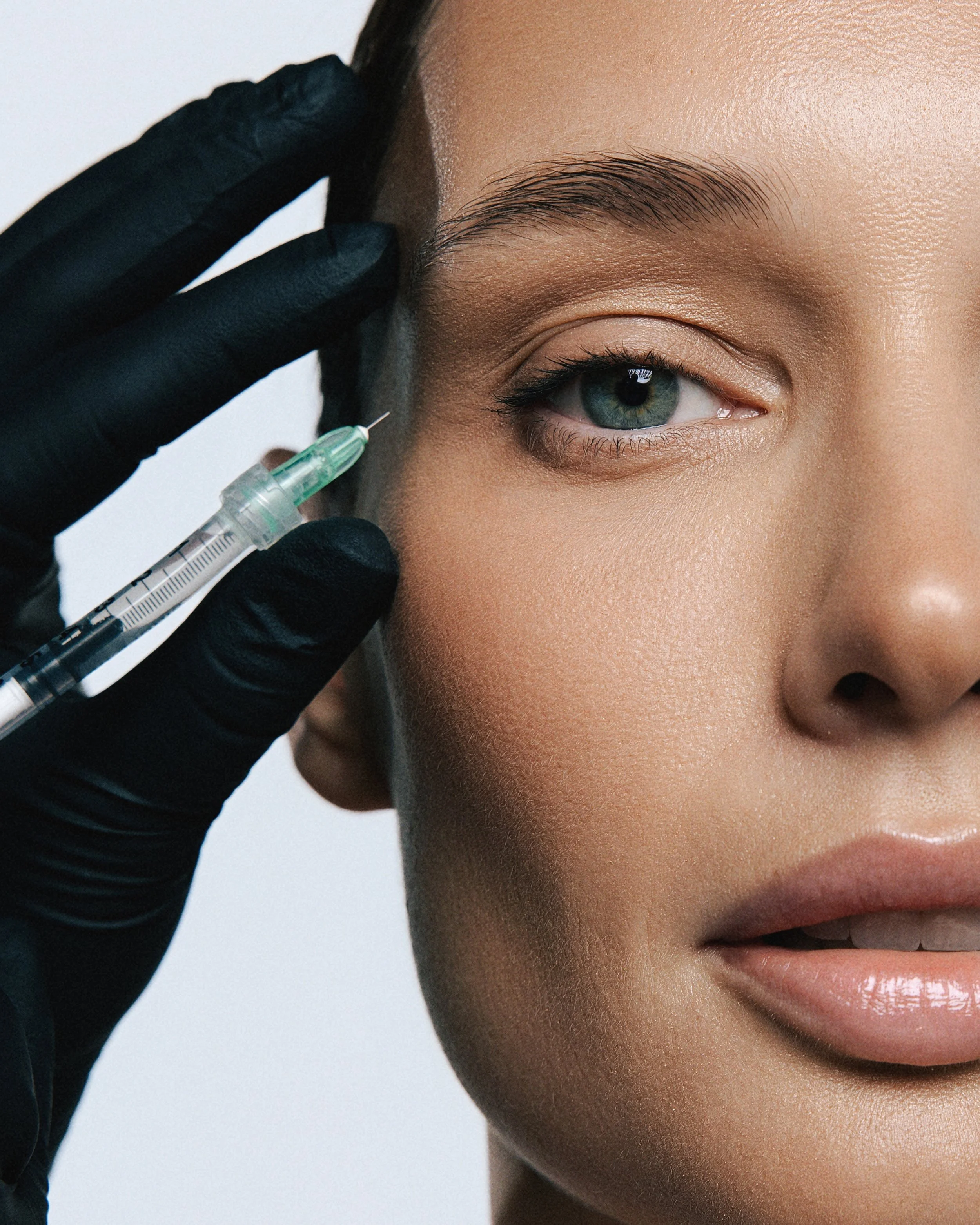 Close-up of a woman receiving a cosmetic injection near her eye, with a gloved hand holding a syringe.