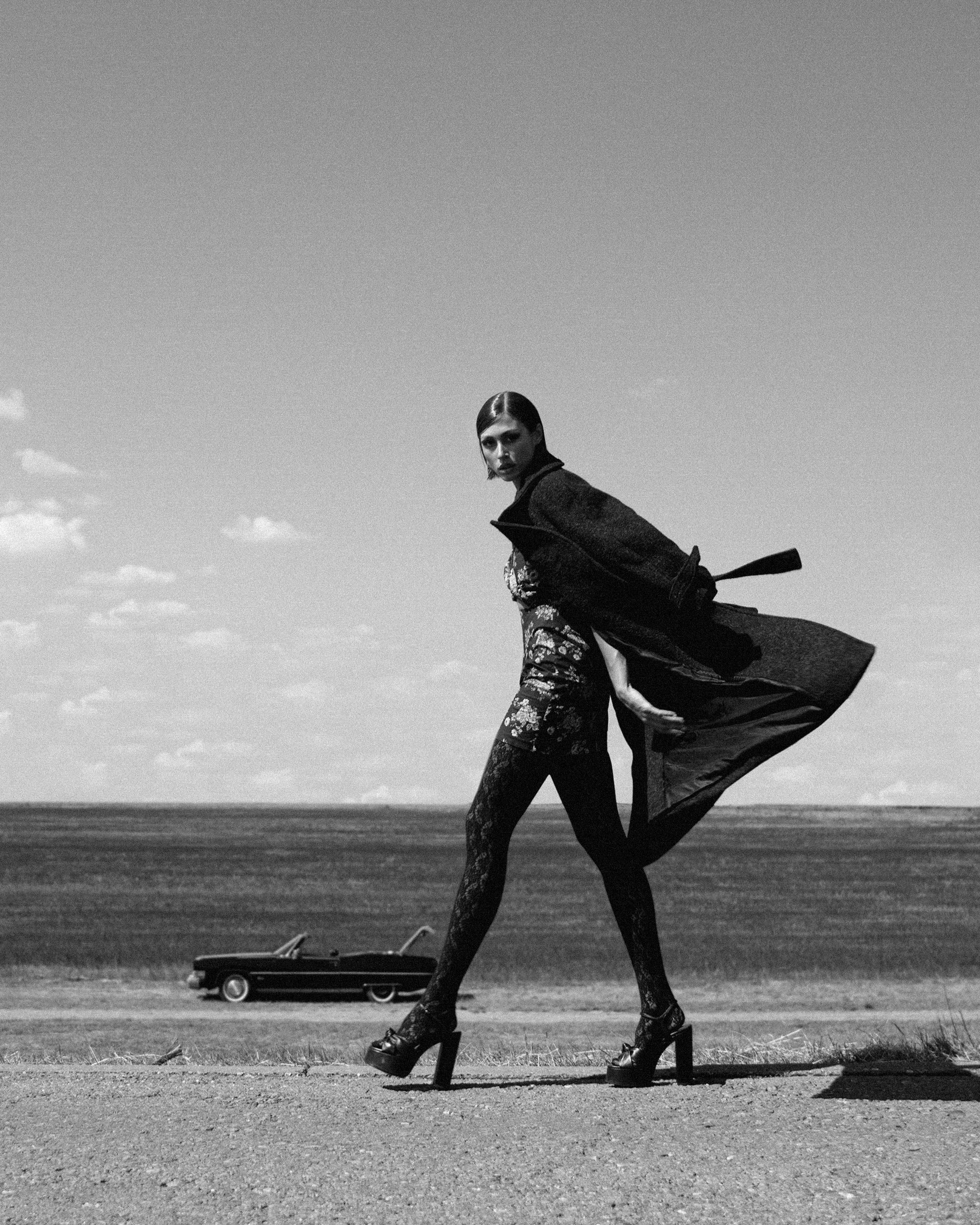 A black-and-white photo of a woman walking on a roadside, wearing high-heeled platform shoes, a patterned jacket, and patterned pants. She has short hair and looks over her shoulder. In the background, there is a vintage car parked on the grass and an open landscape with clouds in the sky.