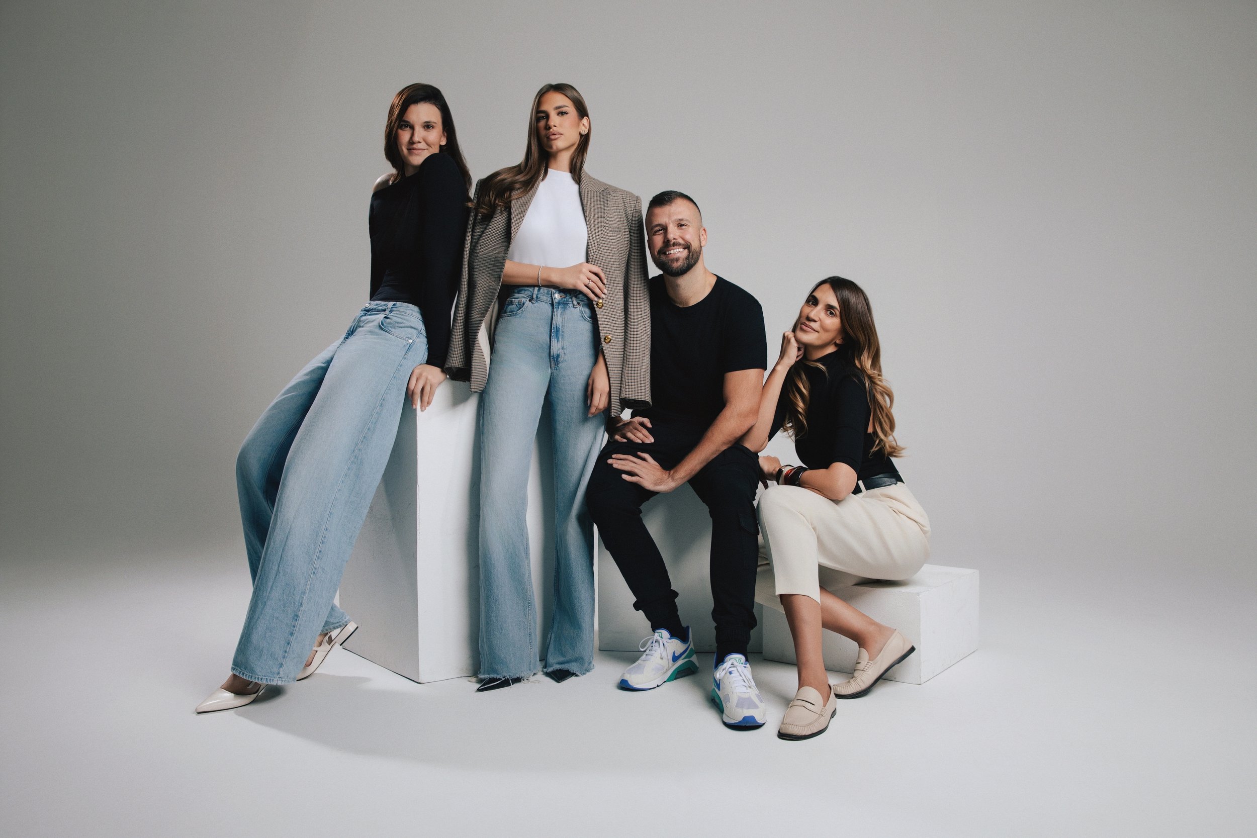 Five young adults posing together against a plain gray background, with some sitting and others standing, dressed in casual and trendy clothing, smiling or with confident expressions.