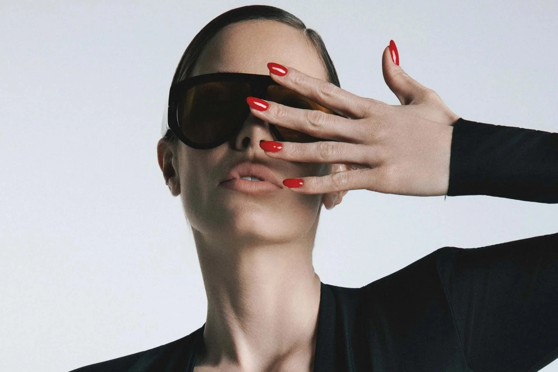 A woman with dark hair and red nail polish covering one eye with her hand, wearing large dark sunglasses and a black top, against a plain light background.