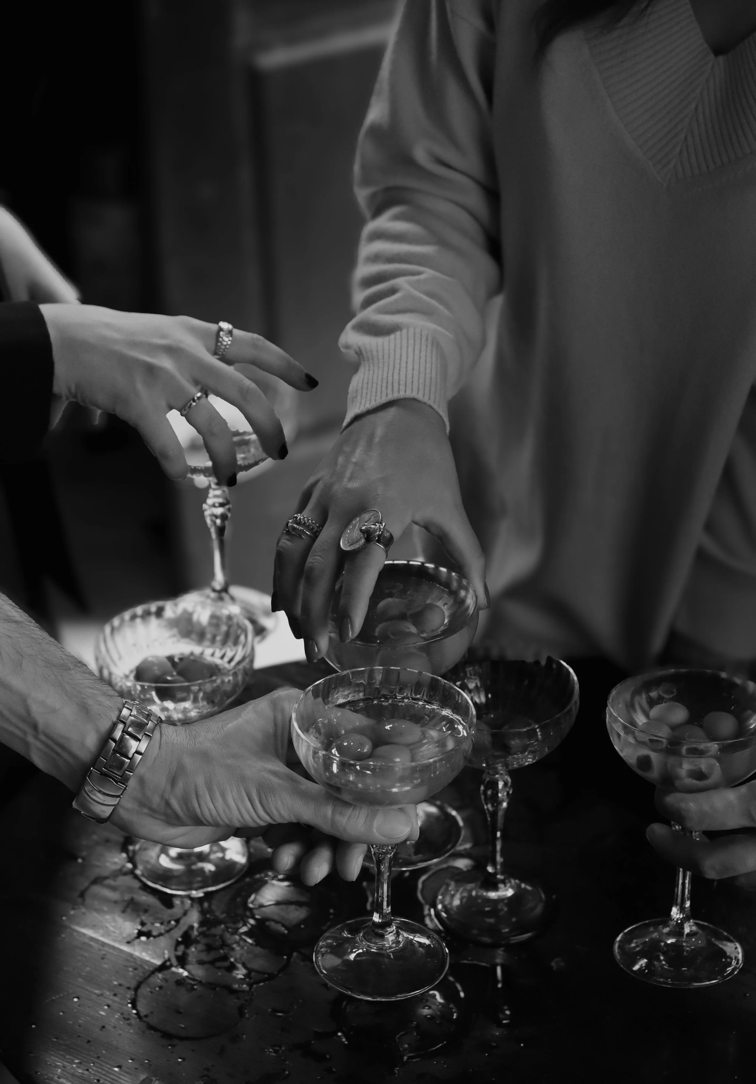 Multiple hands reaching into glass bowls filled with small objects, possibly candies or jewelry, on a wet surface.