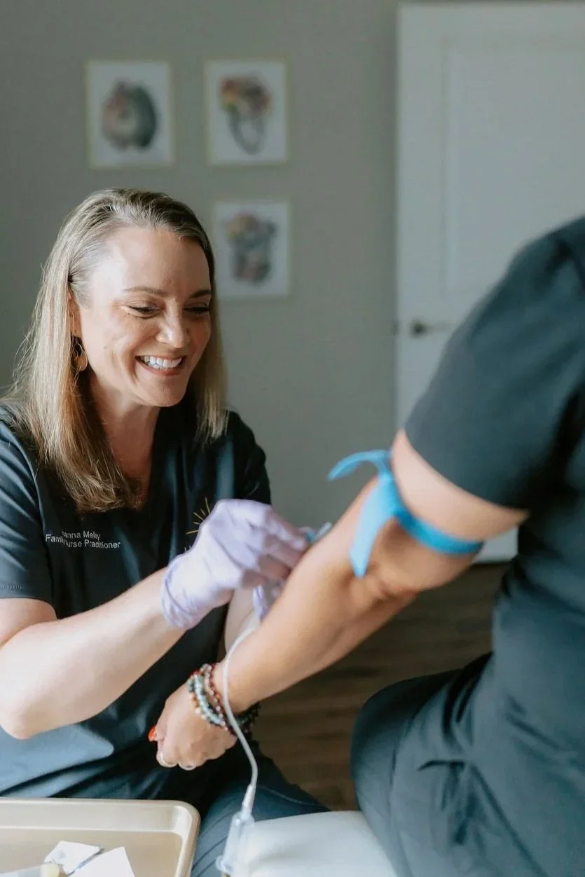 A smiling woman, wearing gloves, is preparing to draw blood from a person using a needle and tube for a medical procedure. The woman appears to be a healthcare professional.