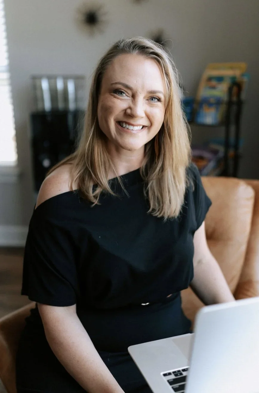 A smiling woman with blonde hair, wearing a black off-shoulder top, sitting in front of a laptop in a cozy living room.