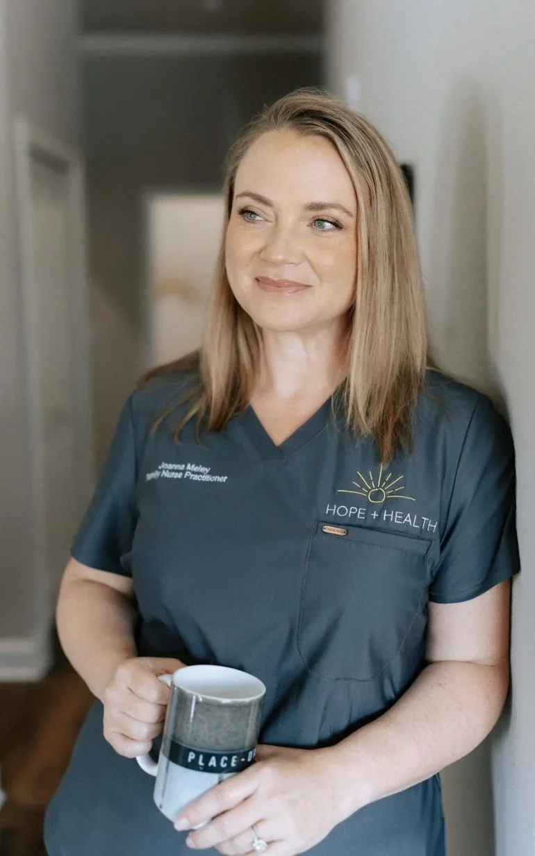 A woman in a dark gray medical scrub top with the words "HOPE + HEALTH" and "Joanne Maley, RN Nurse Practitioner" on it, standing in a room, holding a mug, looking slightly to the side with a gentle smile.