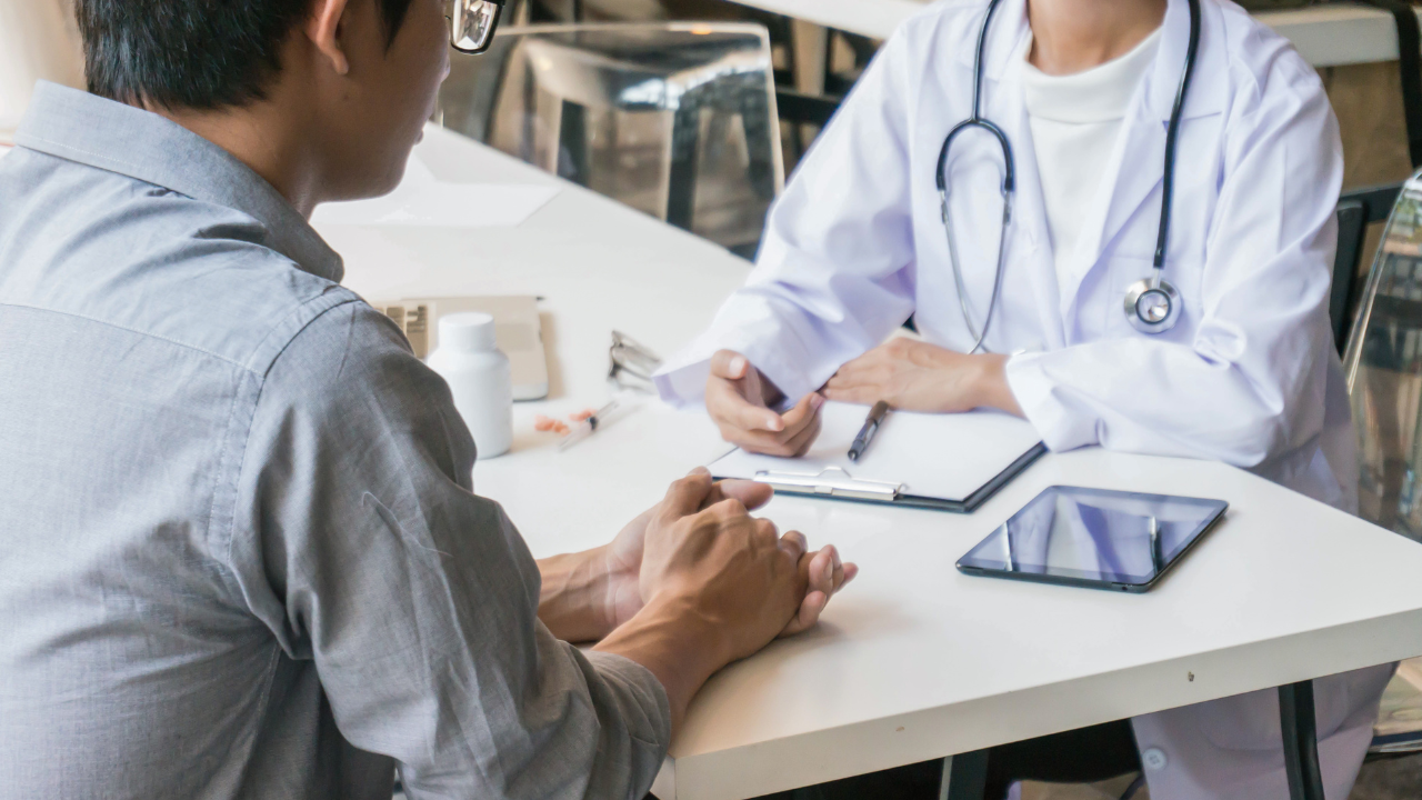 A doctor in a white coat with a stethoscope conversations with a male patient at a table, with medical supplies, a clipboard, and a tablet nearby.