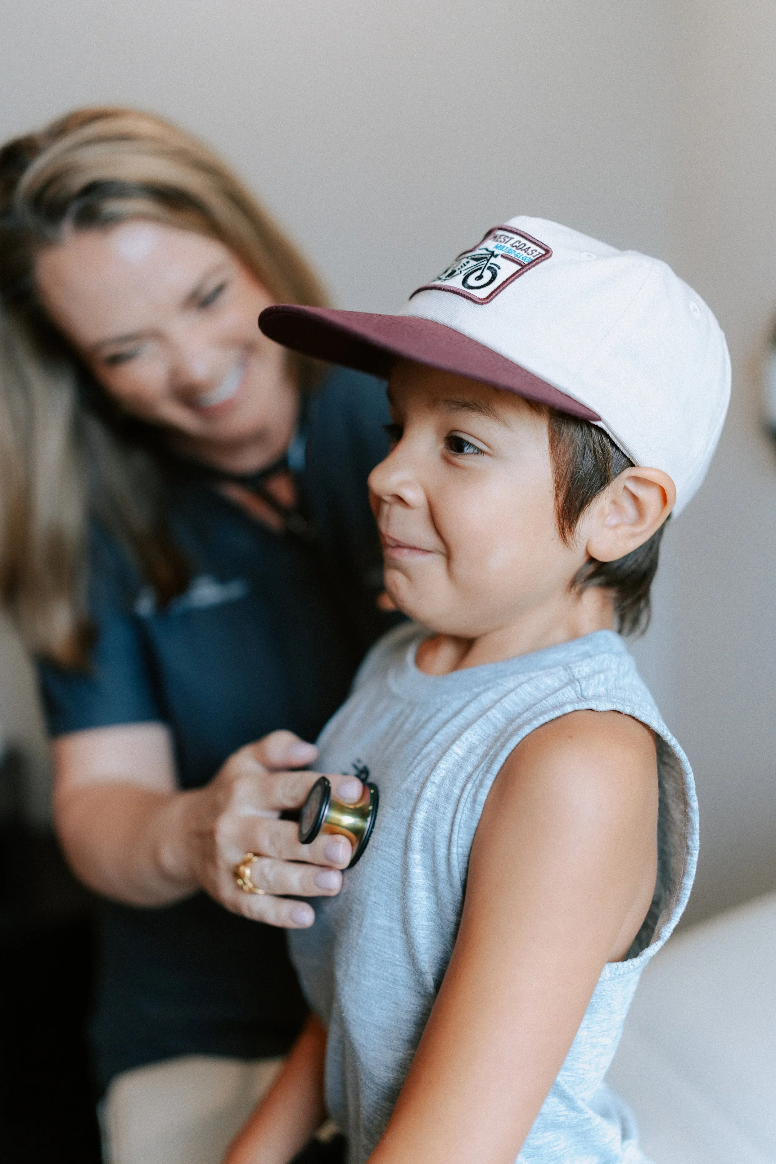 A young boy wearing a white baseball cap with a red brim and a patch. A healthcare professional is examining him with a stethoscope, and they are smiling.