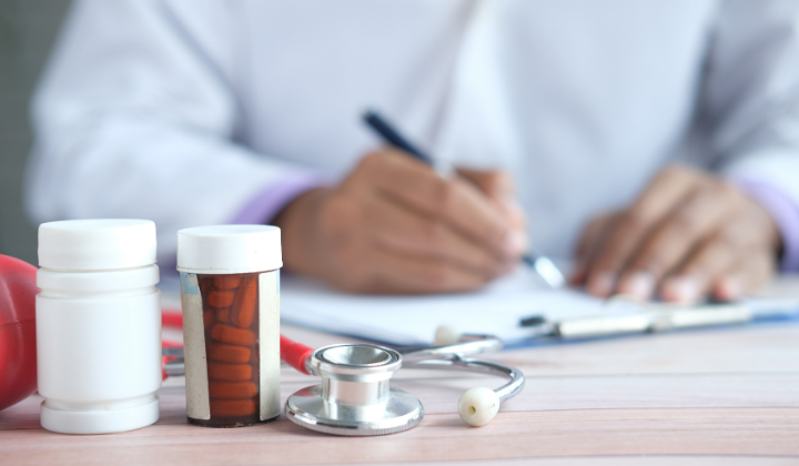 A doctor or medical professional writing in a notebook with medical supplies, including pills, a pill bottle, a stethoscope, and an apple, on a wooden table.