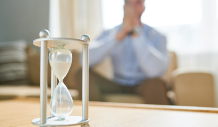 An hourglass in the foreground and a person sitting with hands clasped in the background.