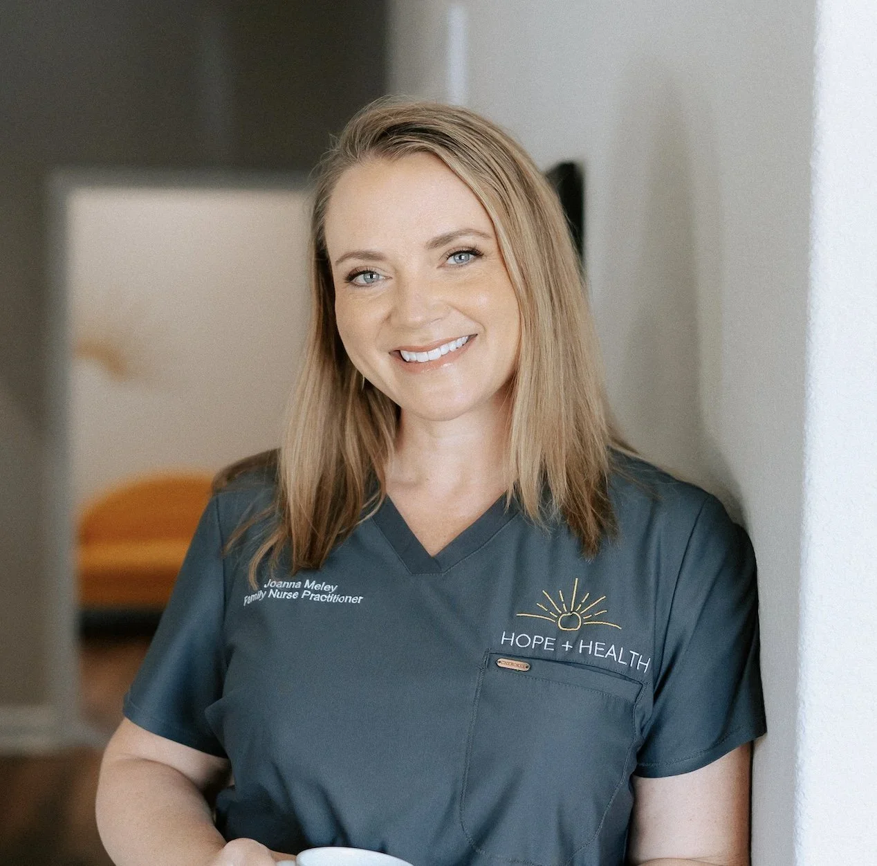 A smiling woman with light brown hair, wearing a dark teal medical scrub top with the text 'Joanna Meloy, Family Nurse Practitioner' and a logo that says 'Hope + Health.' She is standing indoors, leaning against a white wall, with a blurred background that includes a doorway and a piece of furniture.