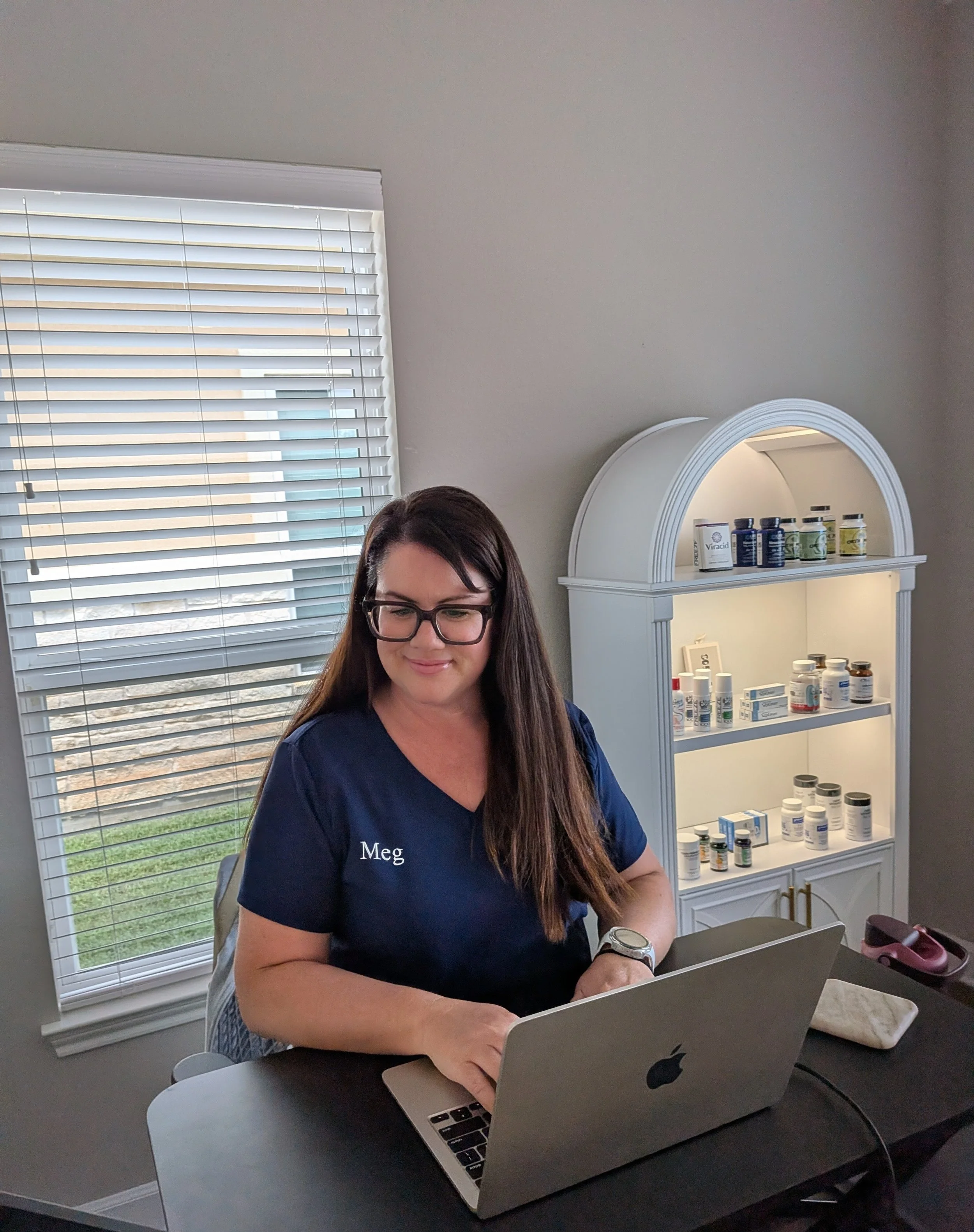 A woman with dark hair and glasses, wearing a navy blue shirt with the name 'Meg' on it, working on a laptop at a black desk in a well-lit room with a window that has blinds and a white cabinet filled with bottles of medication or supplements.