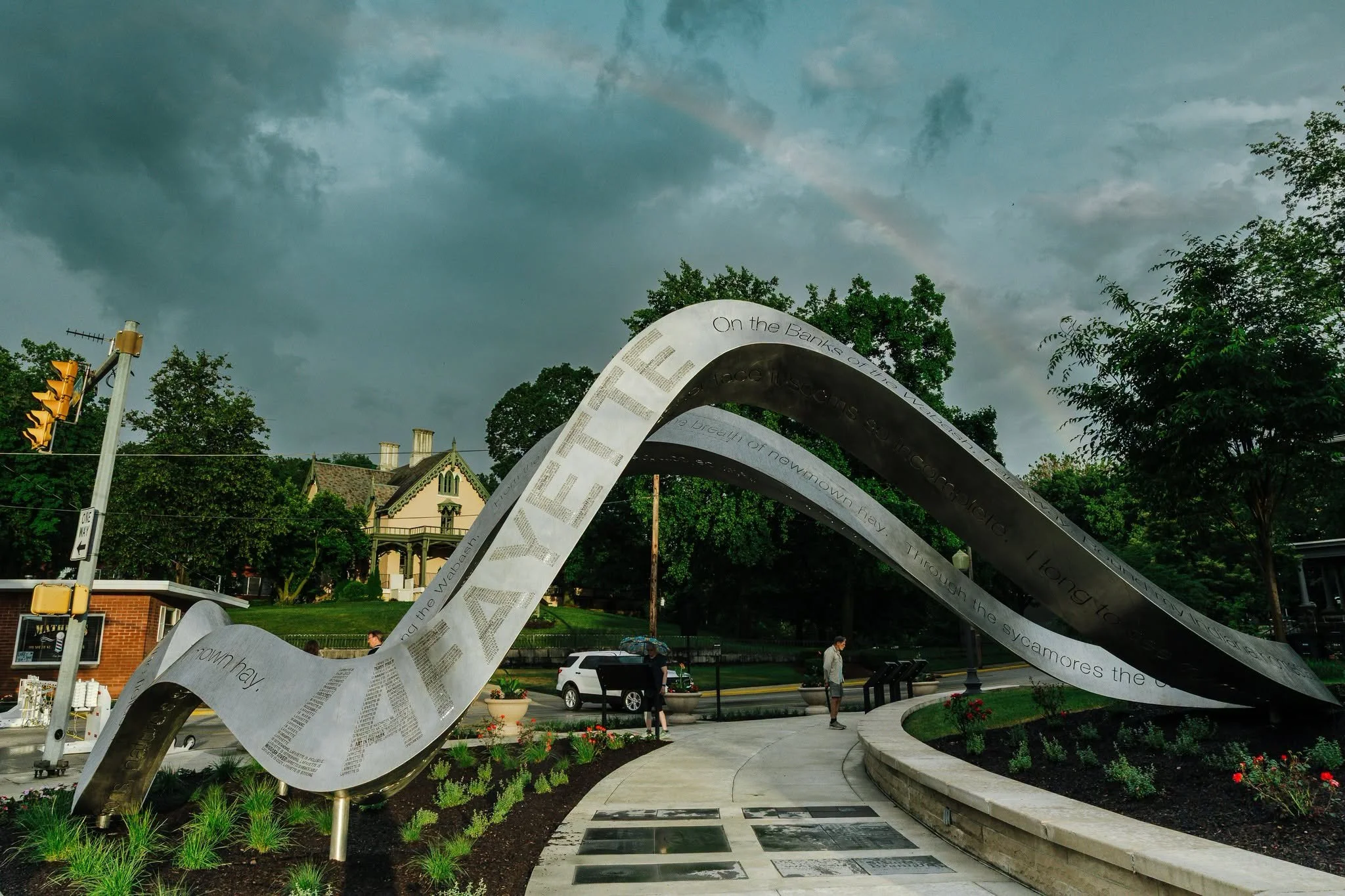 A public park features a metallic ribbon sculpture with engraved text, surrounded by benches, flowers, trees, and a pathway, with buildings, a parked white car, and overcast sky with dark clouds in the background.