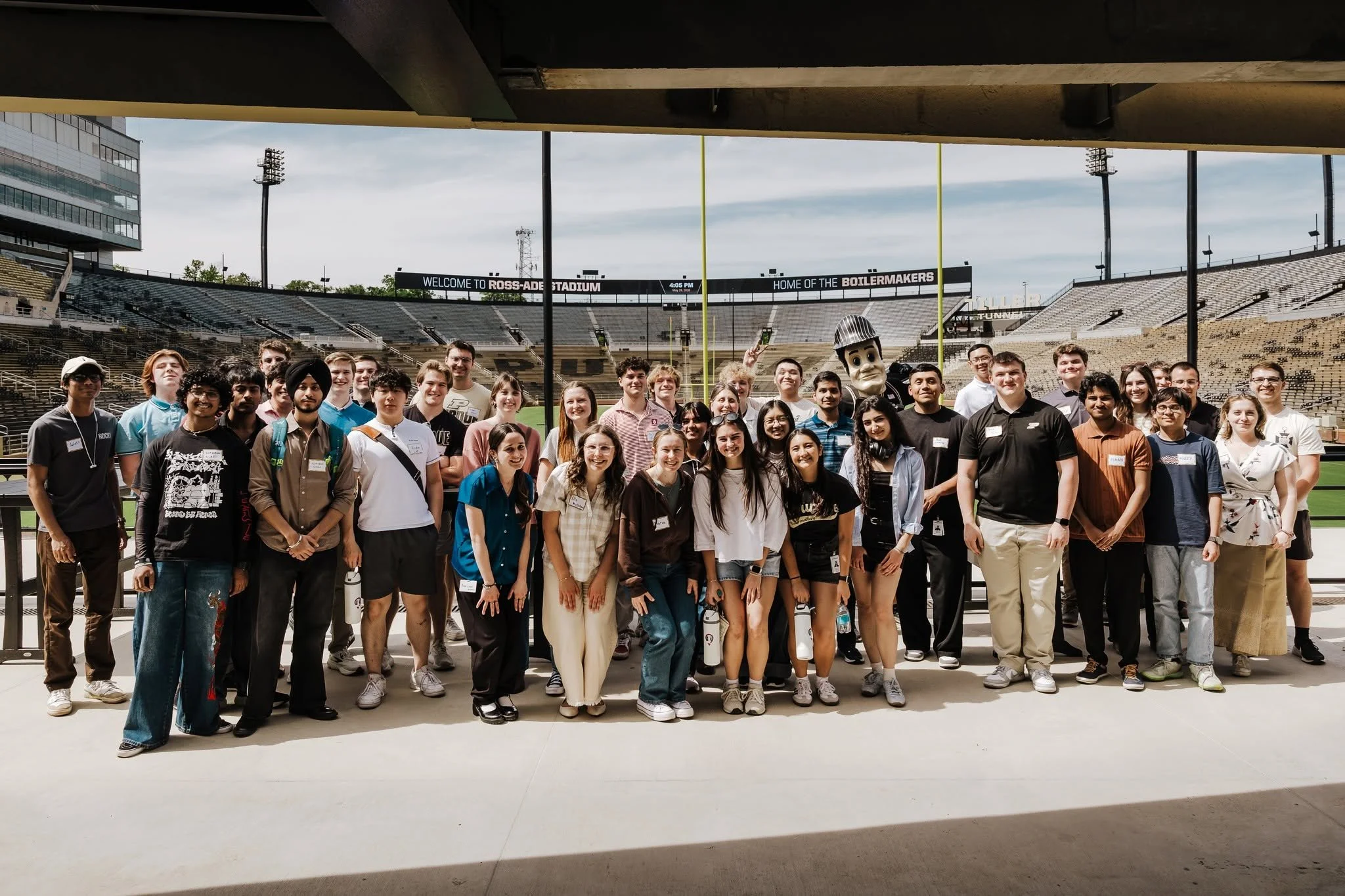 Group of diverse people standing together on a football field in the stadium, smiling for the photo, with the stadium seating and scoreboard in the background.