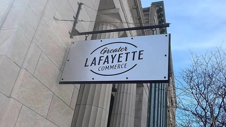 A white rectangular sign with black text reading "Greater Lafayette Commerce" hanging on the side of a building with columns, against a partly cloudy sky and bare tree branches.