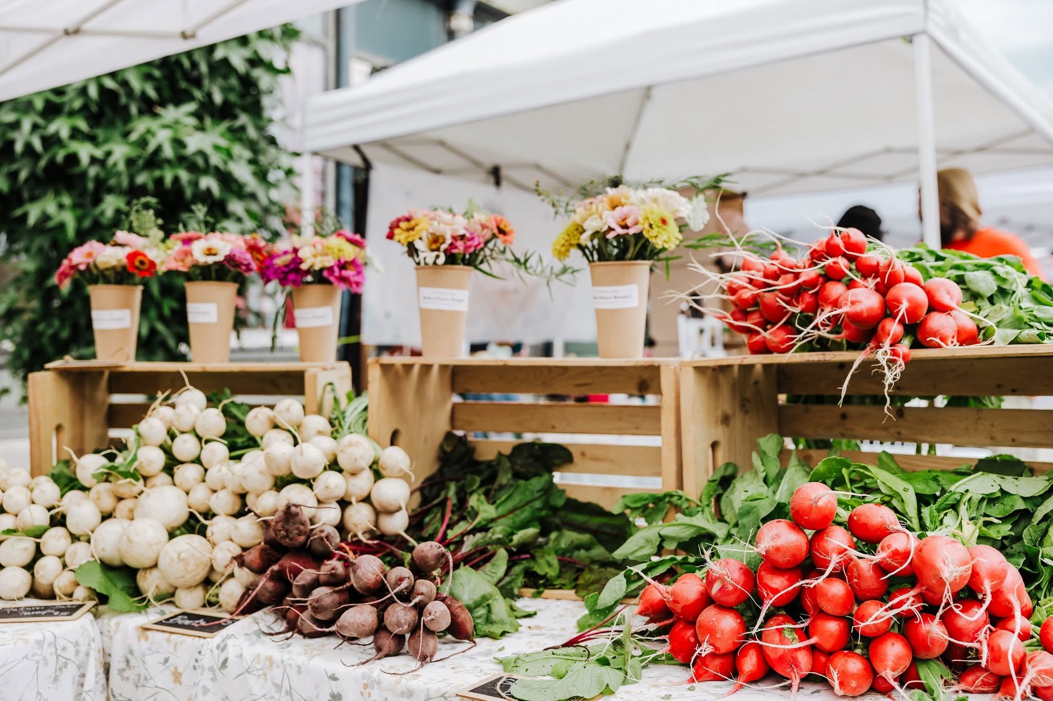 Market produce stand with fresh vegetables and flowers. Front of the stand displays onions and radishes, with flowers in separate containers behind them.