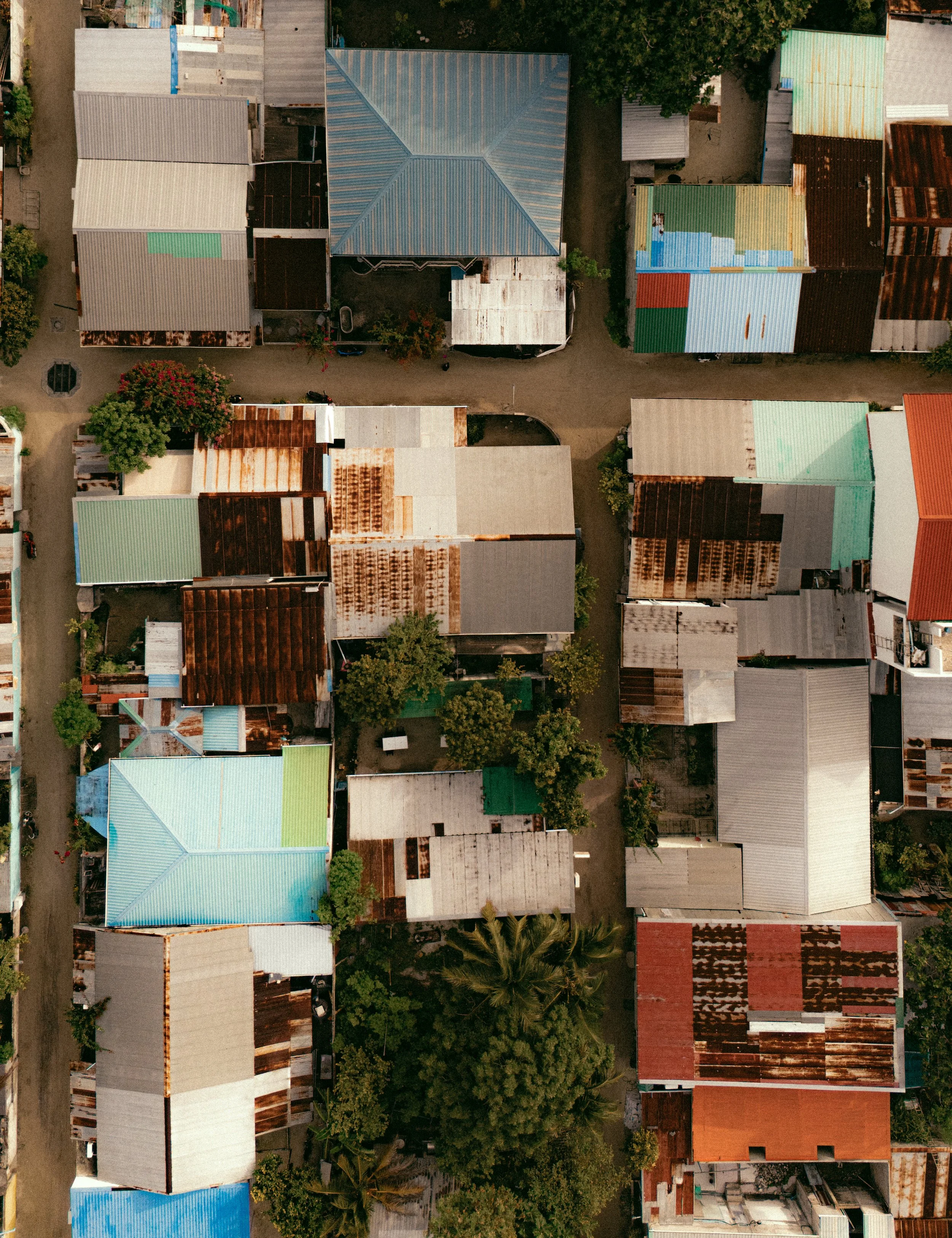 Drone view of the Guraidhoo neighborhood with houses featuring colorful roofs, trees, and dirt paths between buildings, the tucked away side of Maldives.