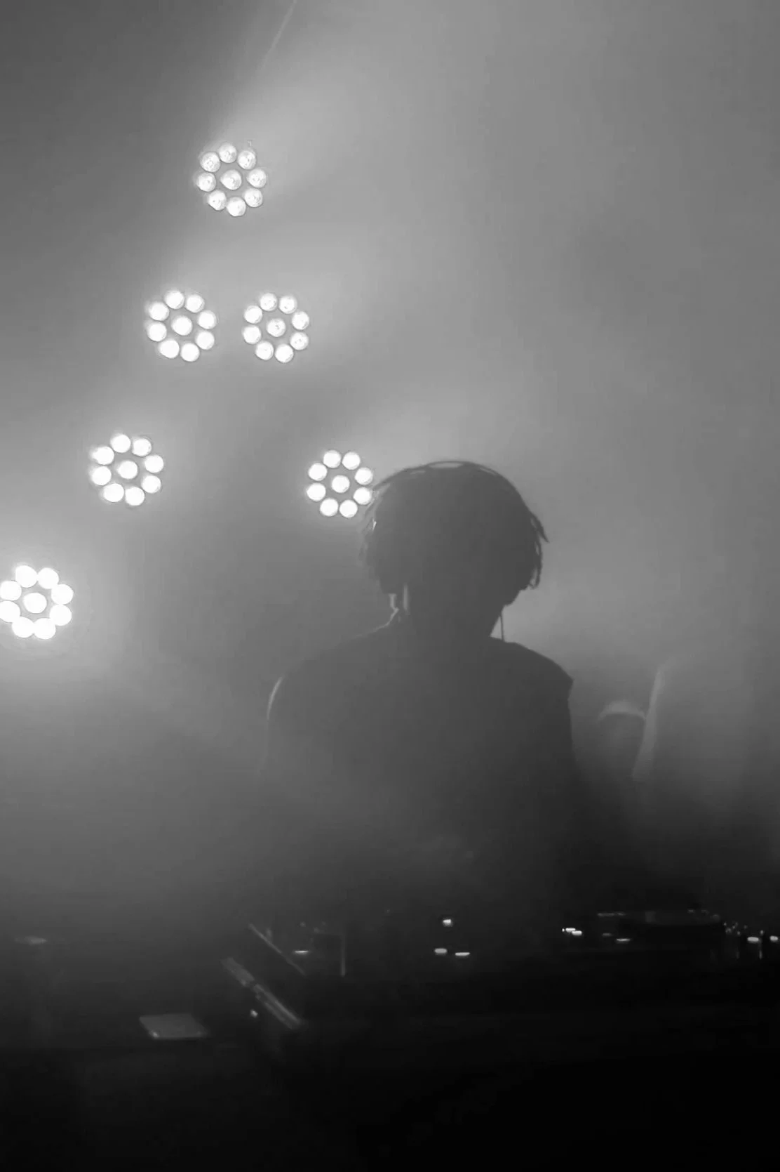 Silhouette d'une personne avec des écouteurs, en arrière-plan des lumières de scène ou de DJ dans une ambiance de concert ou de club, en noir et blanc.