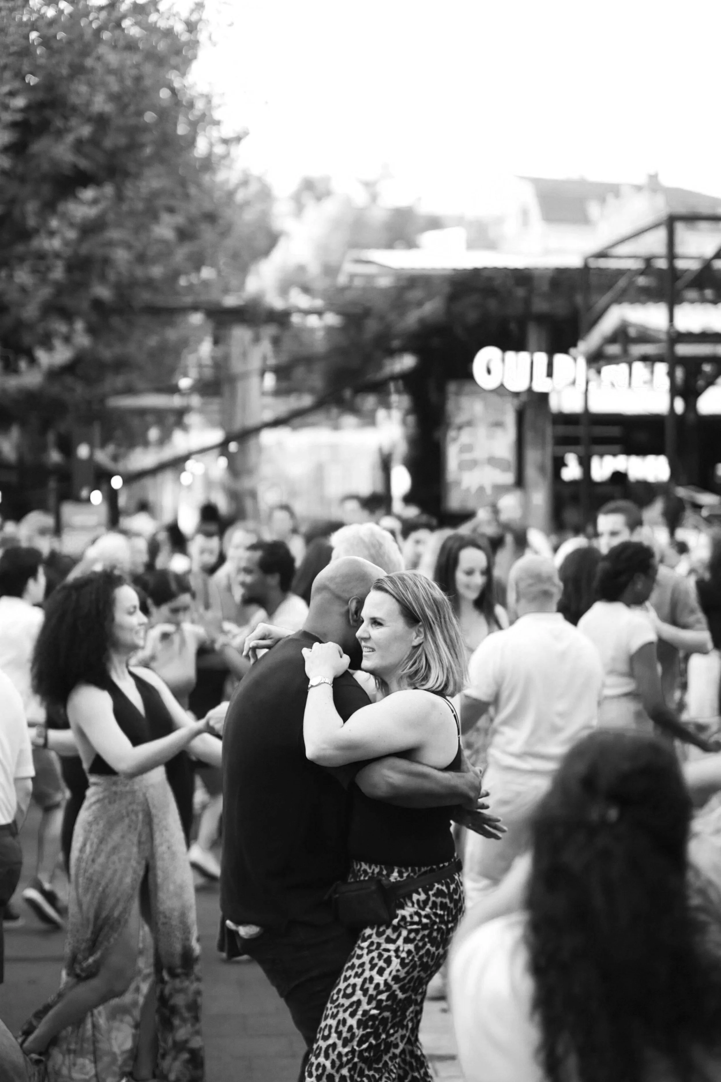 Une scène de danse en plein air avec plusieurs couples qui dansent, un public regardant et des bâtiments en arrière-plan, image en noir et blanc.