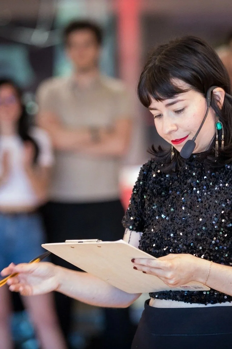 Une femme portant un haut noir brillant avec un casque de communication, tenant un carnet et un stylo, semble donner des instructions ou faire une annonce lors d'un événement ou d'une présentation.