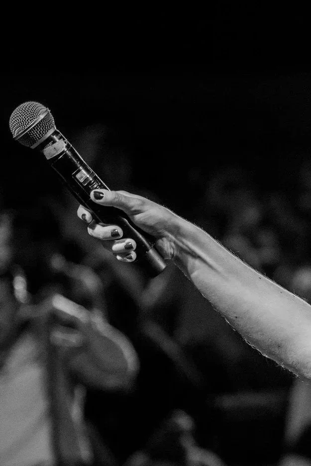 Une main tenant un microphone lors d'une performance ou d'un discours, fond flou avec une audience