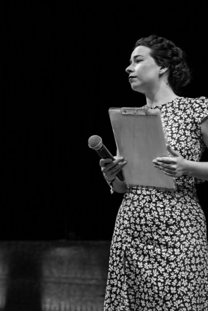 Une femme debout, tenant un micro et un dossier, regardant de côté, en noir et blanc