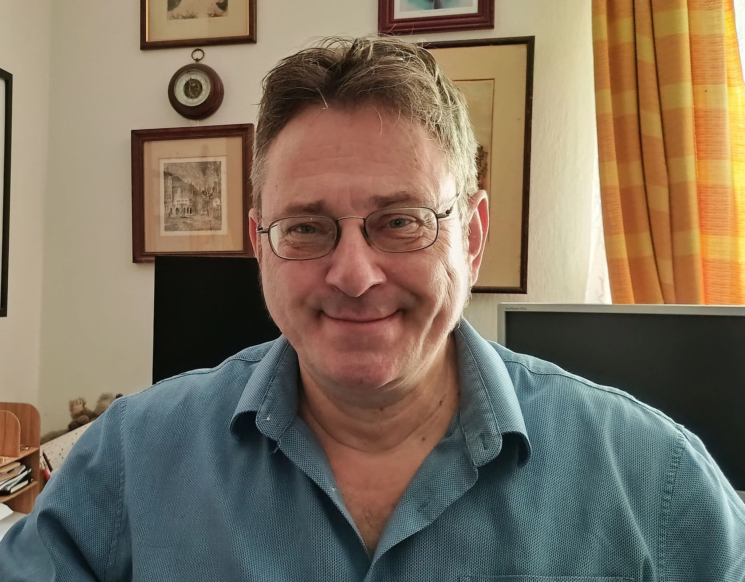 A smiling man with glasses sits in a room with framed artwork on the wall behind him and a curtain to his right.