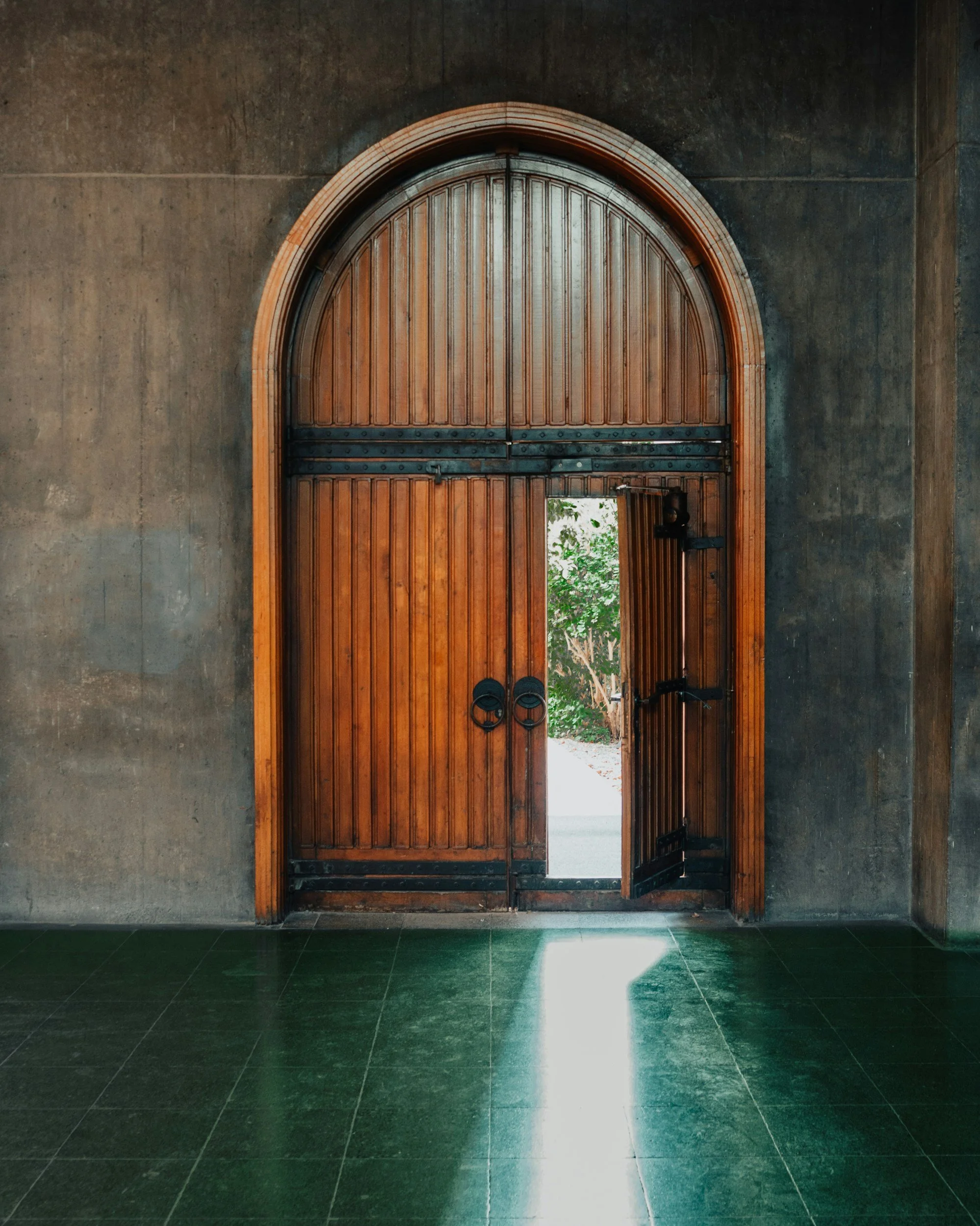 A large wooden arched double door set into a concrete wall, with metal hinges and a latch. The door is partially open, revealing greenery outside. Green tile flooring is visible in the foreground, with sunlight creating a bright patch on the floor near the door.