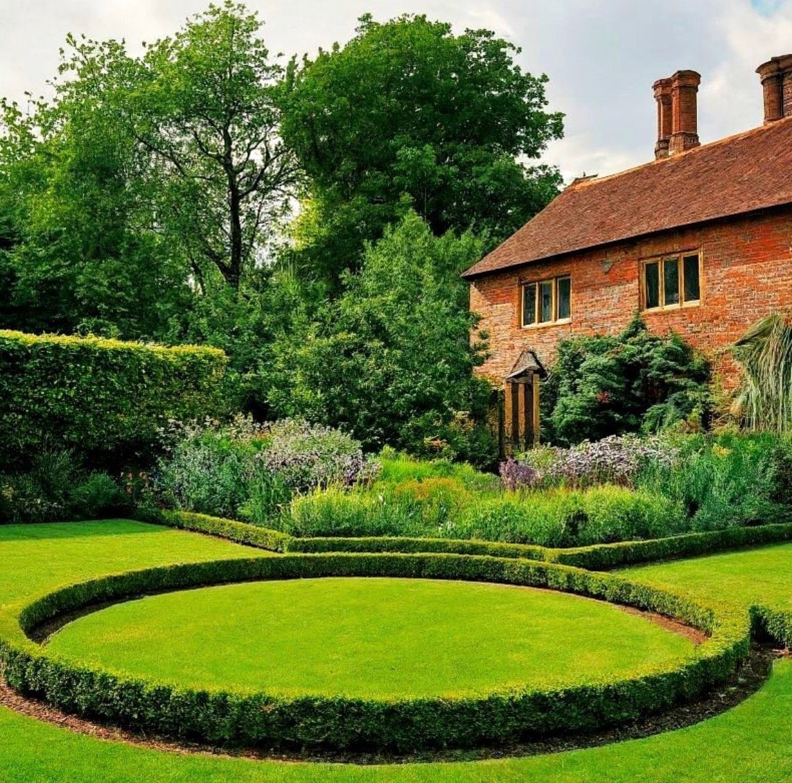 A well-maintained garden with a circular hedge in front of a brick house surrounded by green trees and plants.