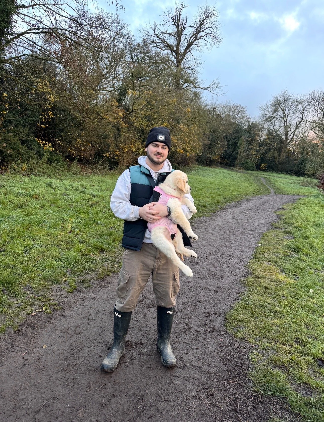 A man standing on a dirt path holding a yellow Labrador puppy in his arms. The man is wearing a black beanie, a gray hoodie, a black vest, beige cargo pants, and tall black rain boots. The background features trees with some autumn foliage and a cloudy sky.