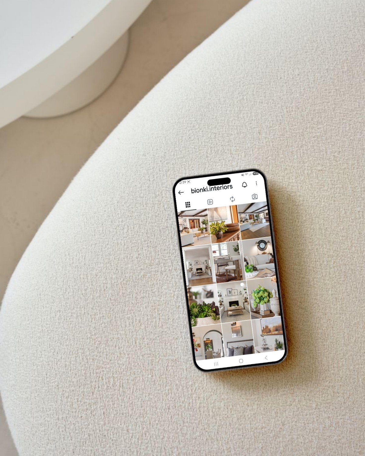A smartphone resting on a beige textured surface displaying an interior design company's Instagram page with photos of modern, stylish home interiors.